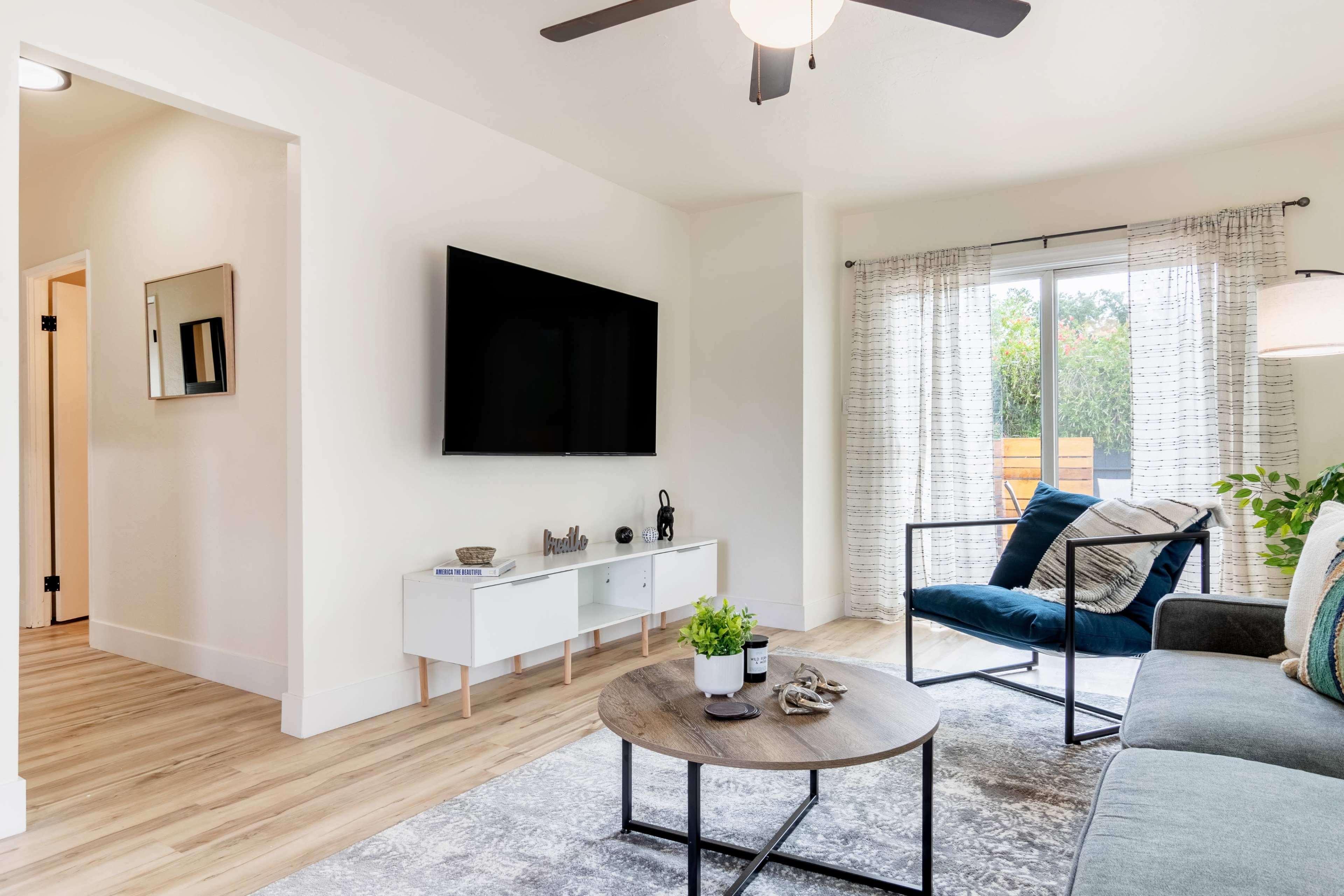The living room features a television mounted on a white wall, a round coffee table, and a blue sofa, with natural light streaming in through large sliding glass doors.