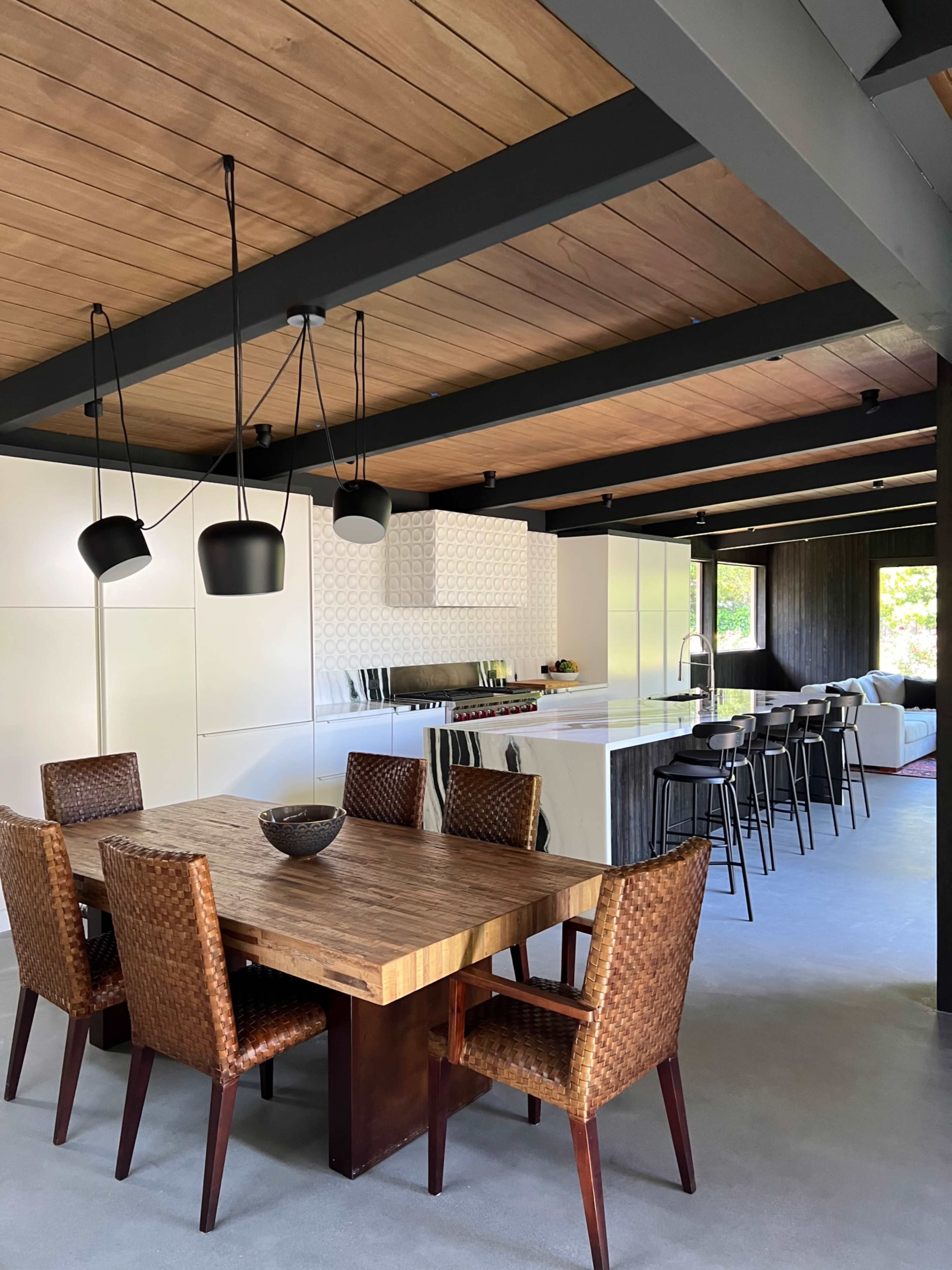 A modern kitchen and dining area with a wooden dining table surrounded by woven chairs, and a sleek kitchen countertop with black bar stools nearby.
