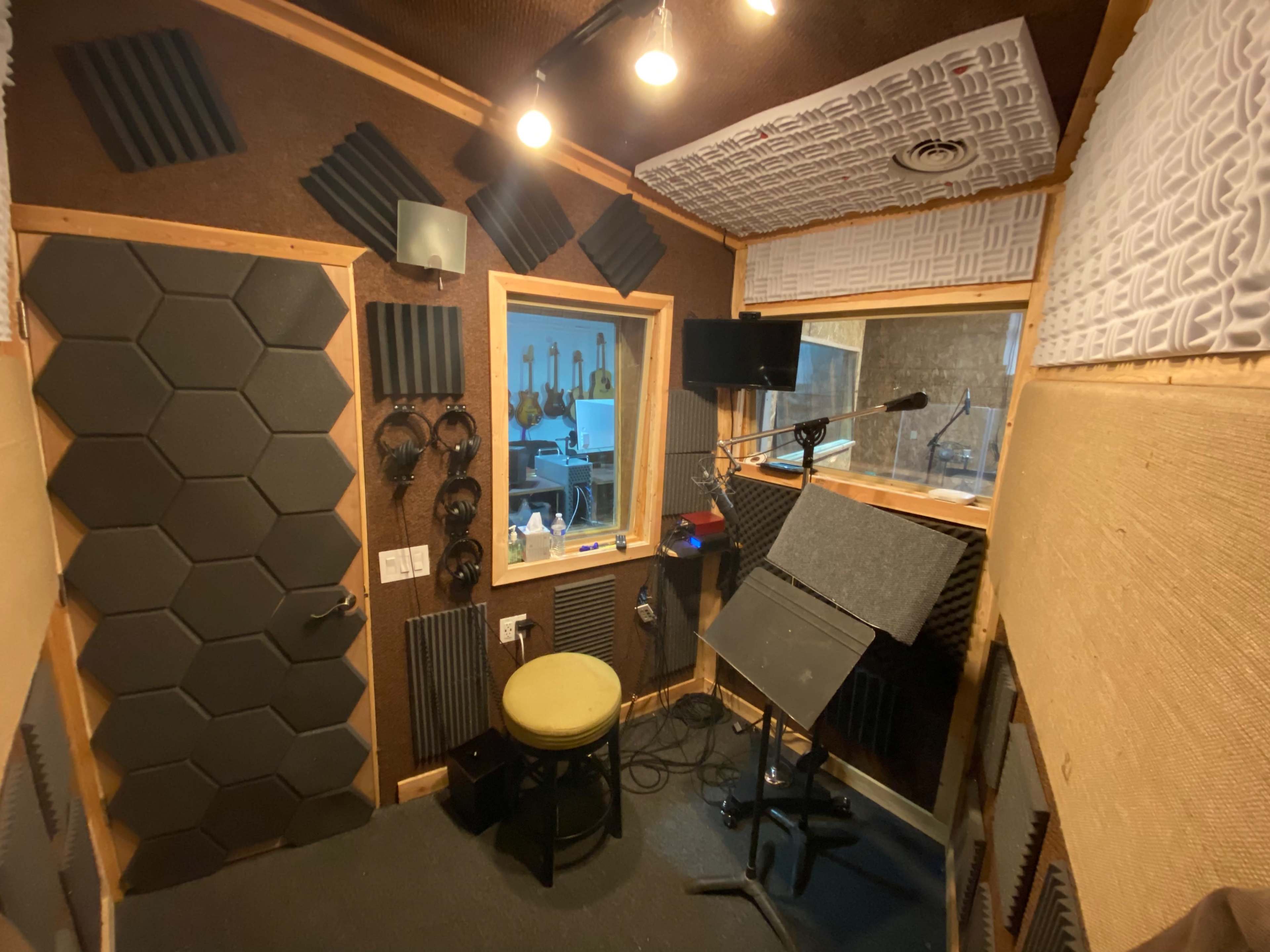 The image shows a soundproof recording studio with acoustic panels, a microphone set up near a music stand, headphones on the wall, and a window overlooking another room.