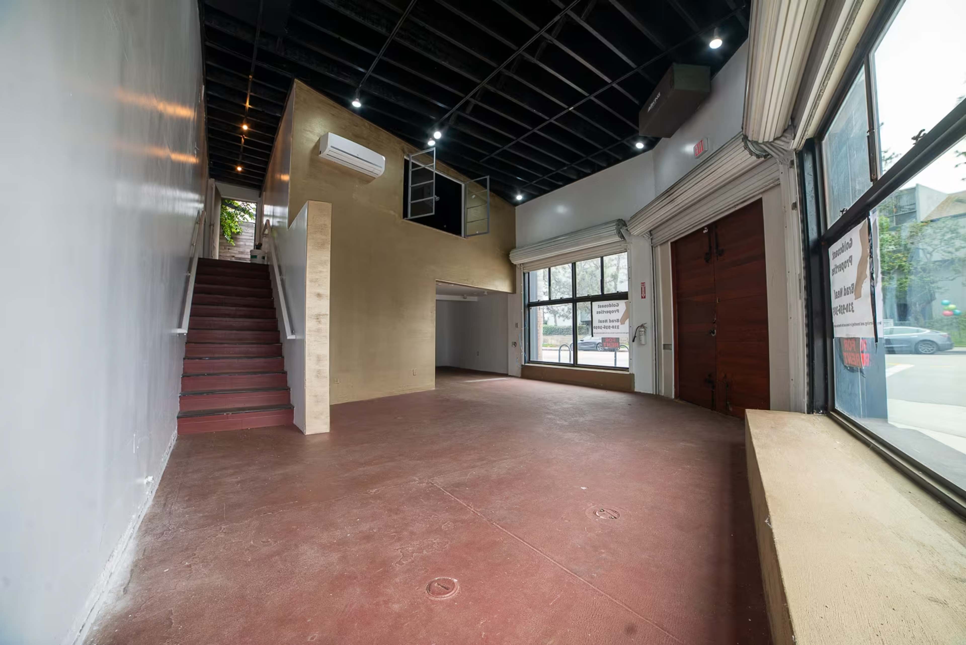 The image shows an empty commercial space with wooden stairs leading to a higher level on the left and large windows facing the street on the right.