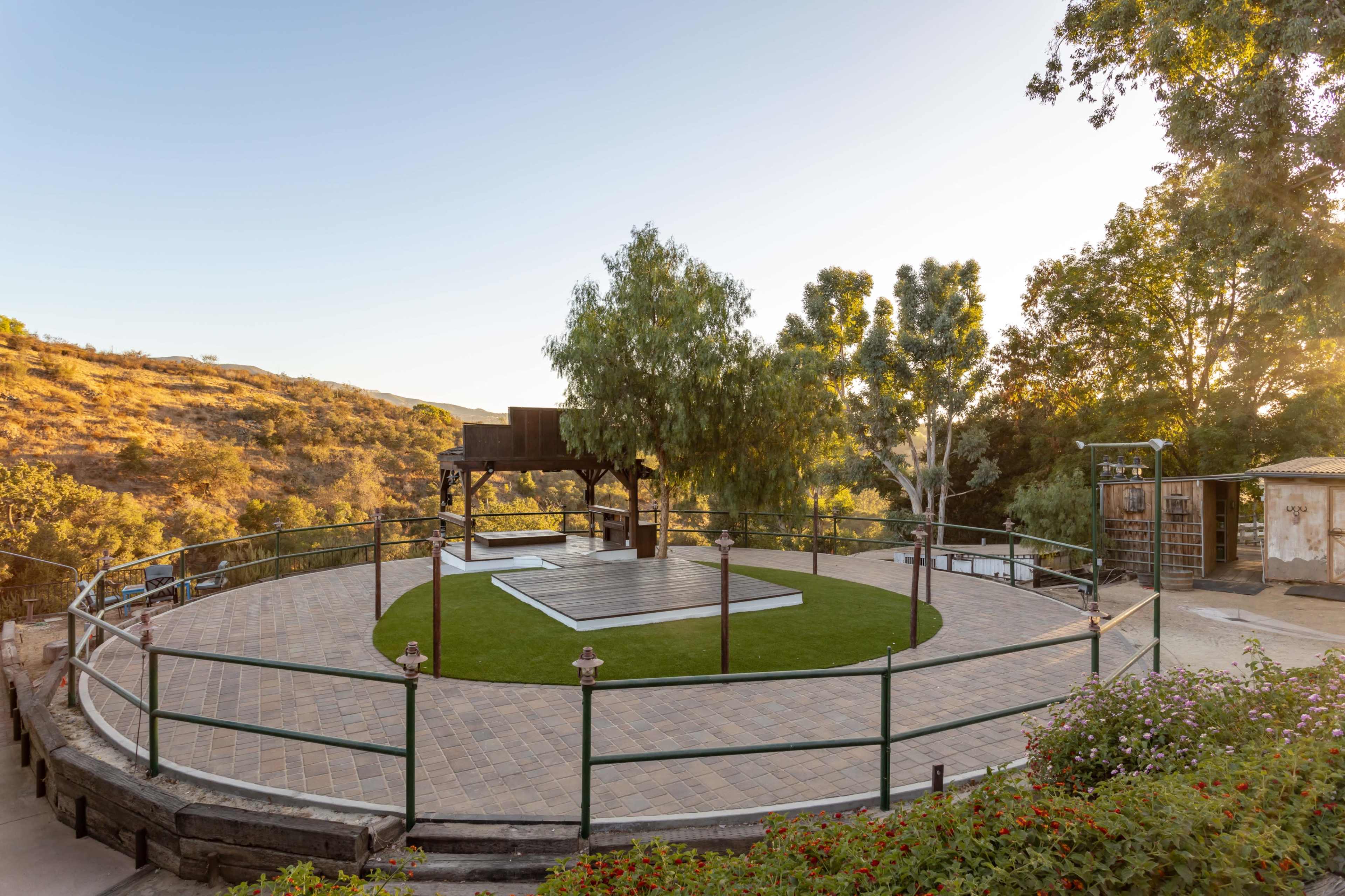 A circular outdoor event space surrounded by greenery, featuring a covered platform and a lawn area.