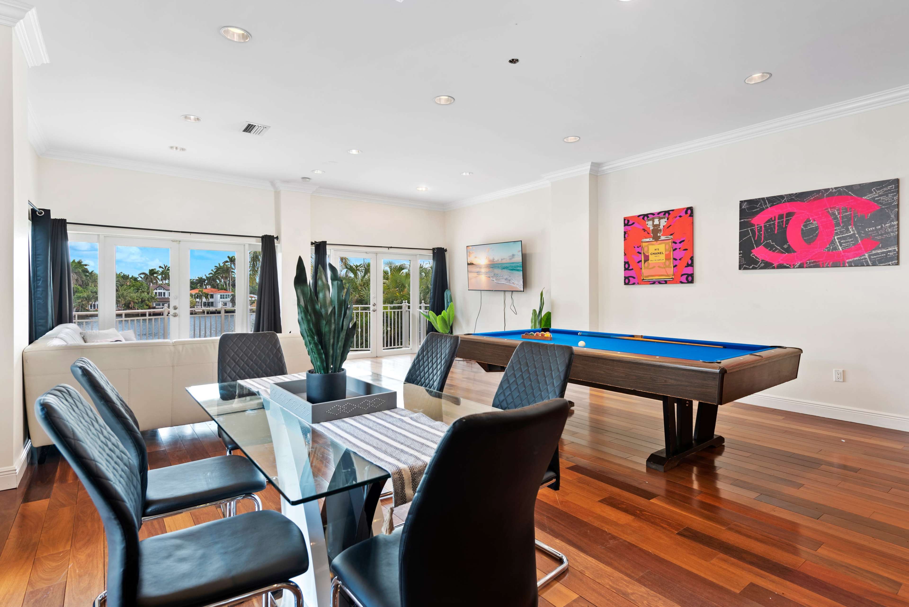 A bright interior space featuring a dining area with a glass table and black chairs, a pool table, and large windows overlooking a balcony.