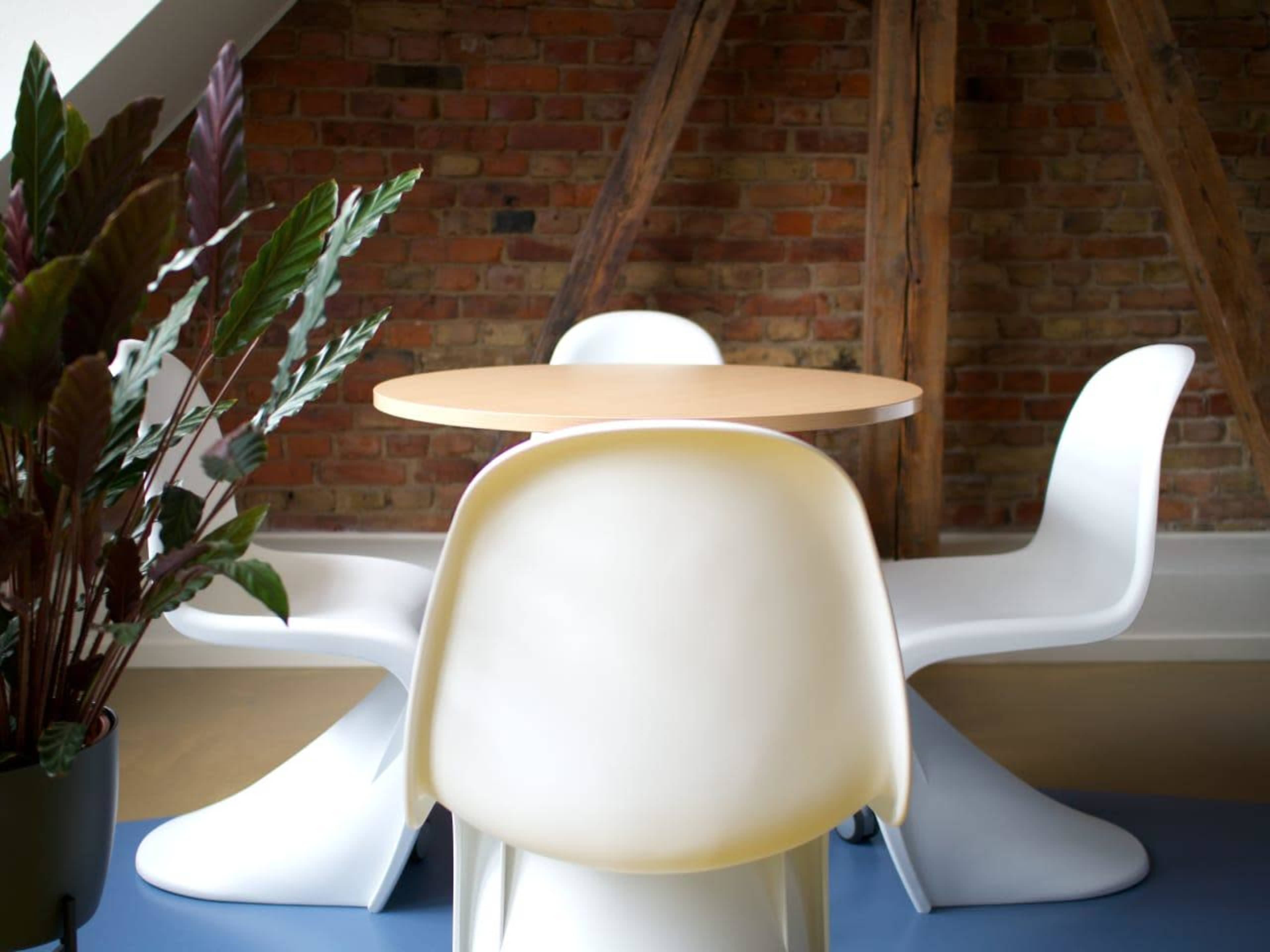 A round wooden table is set in a room with exposed brick walls, accompanied by three white chairs and a potted plant in the foreground.