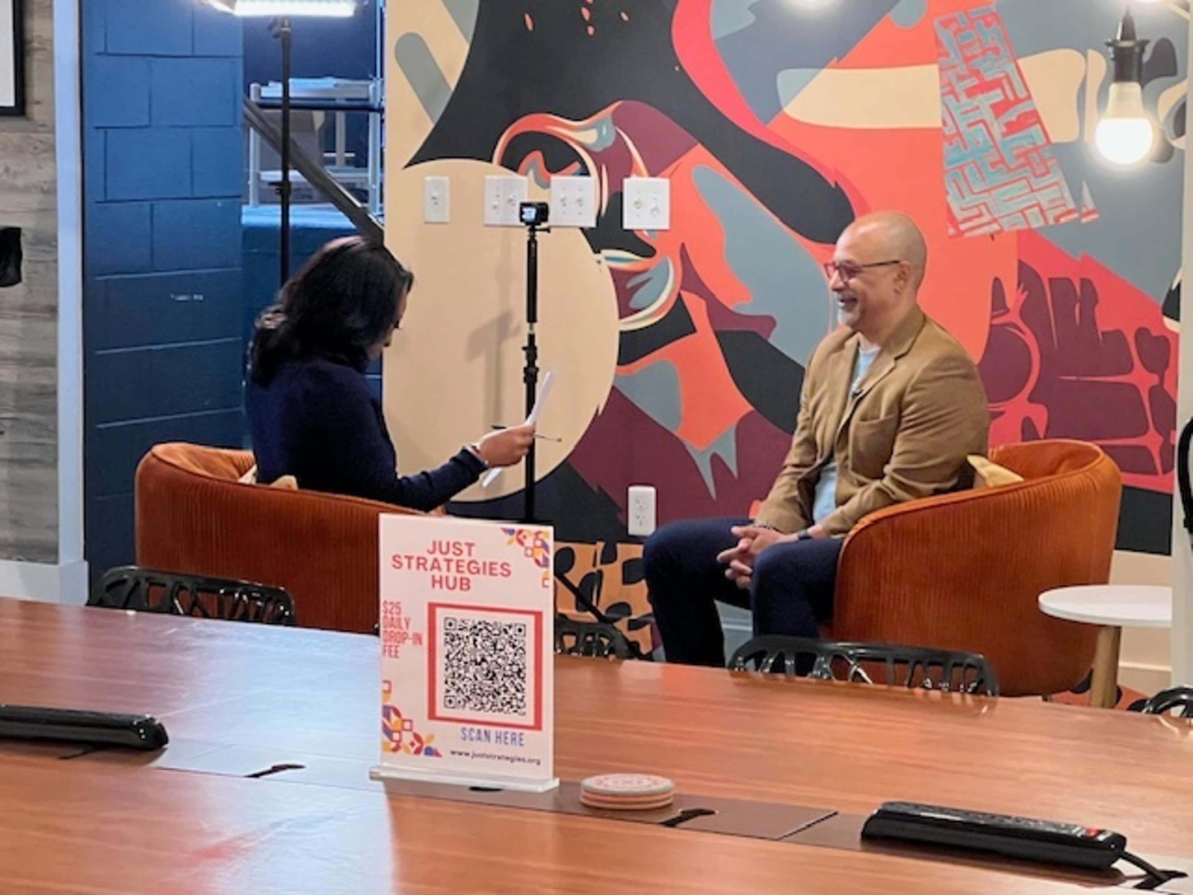 A man and a woman are engaged in an interview while seated on orange chairs in front of a colorful mural, with a camera setup and a QR code sign visible on the table.