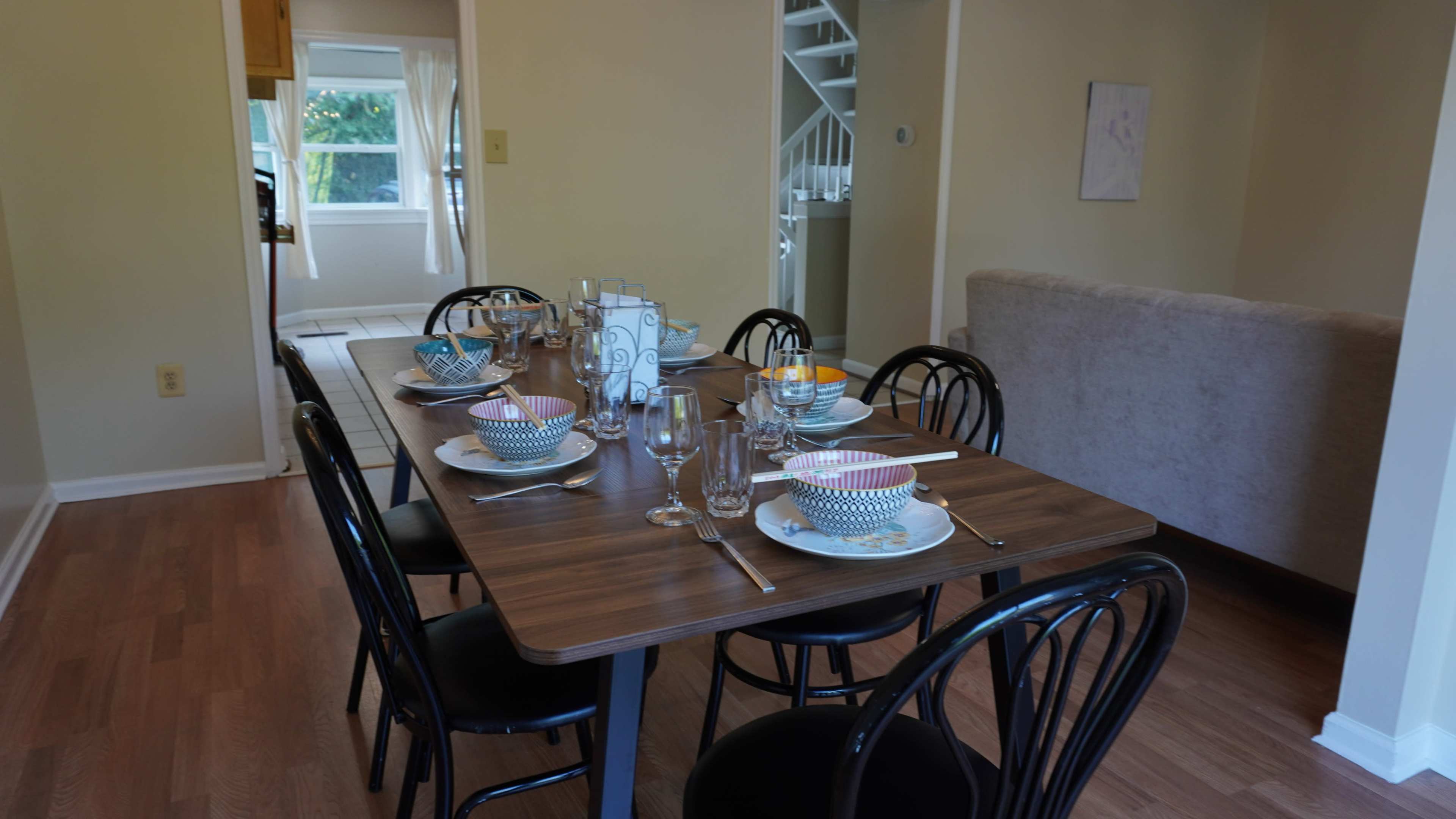 A dining table is set with dishes and glassware, positioned in a well-lit room with a visible kitchen and staircase in the background.