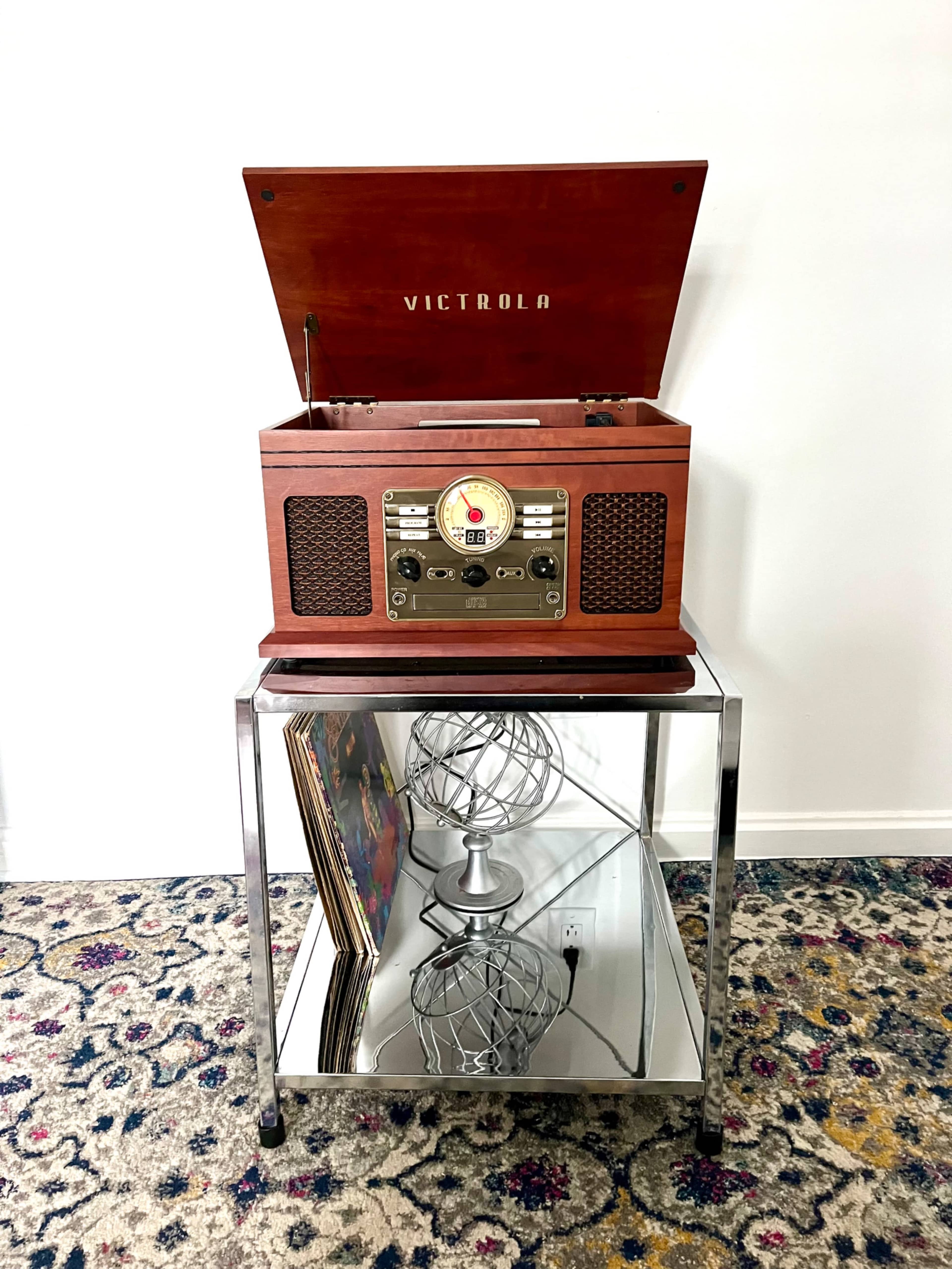 A wooden Victrola record player sits on a chrome table, displaying records and a decorative wire globe below.