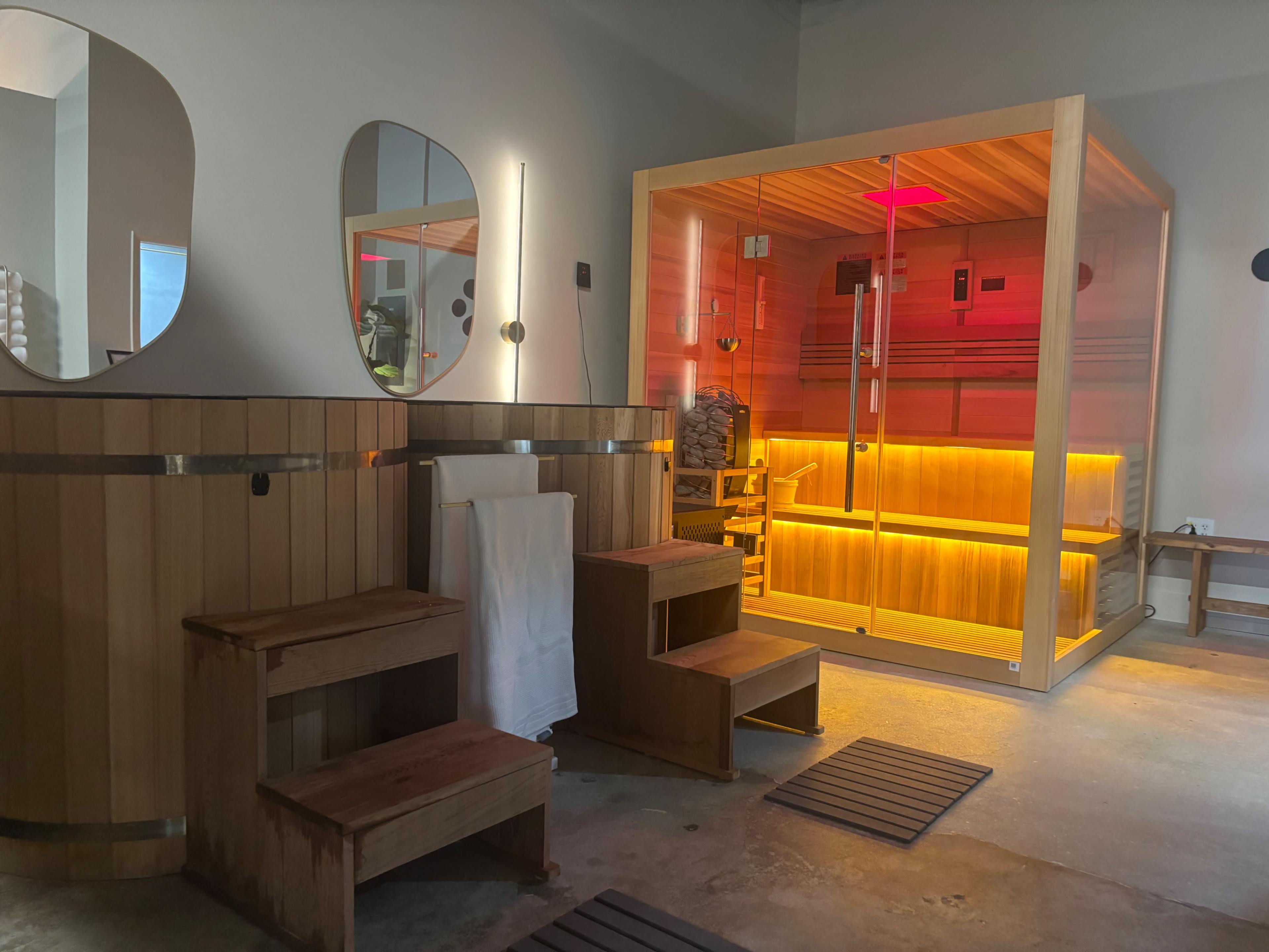 The image shows a modern wellness area featuring a wooden sauna, two mirrors on the wall, and wooden steps leading to the sauna.