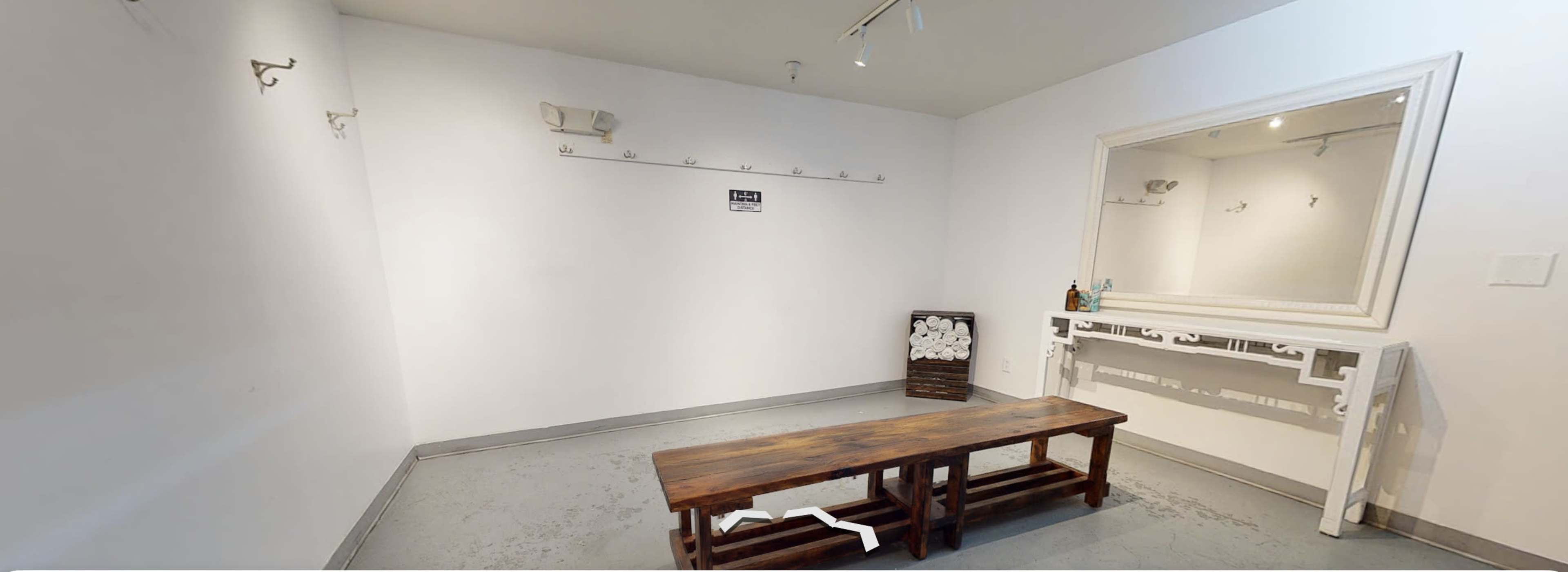 A small, minimalist room featuring a wooden bench, a mirror, hooks on the walls, and a shelf with toiletries.
