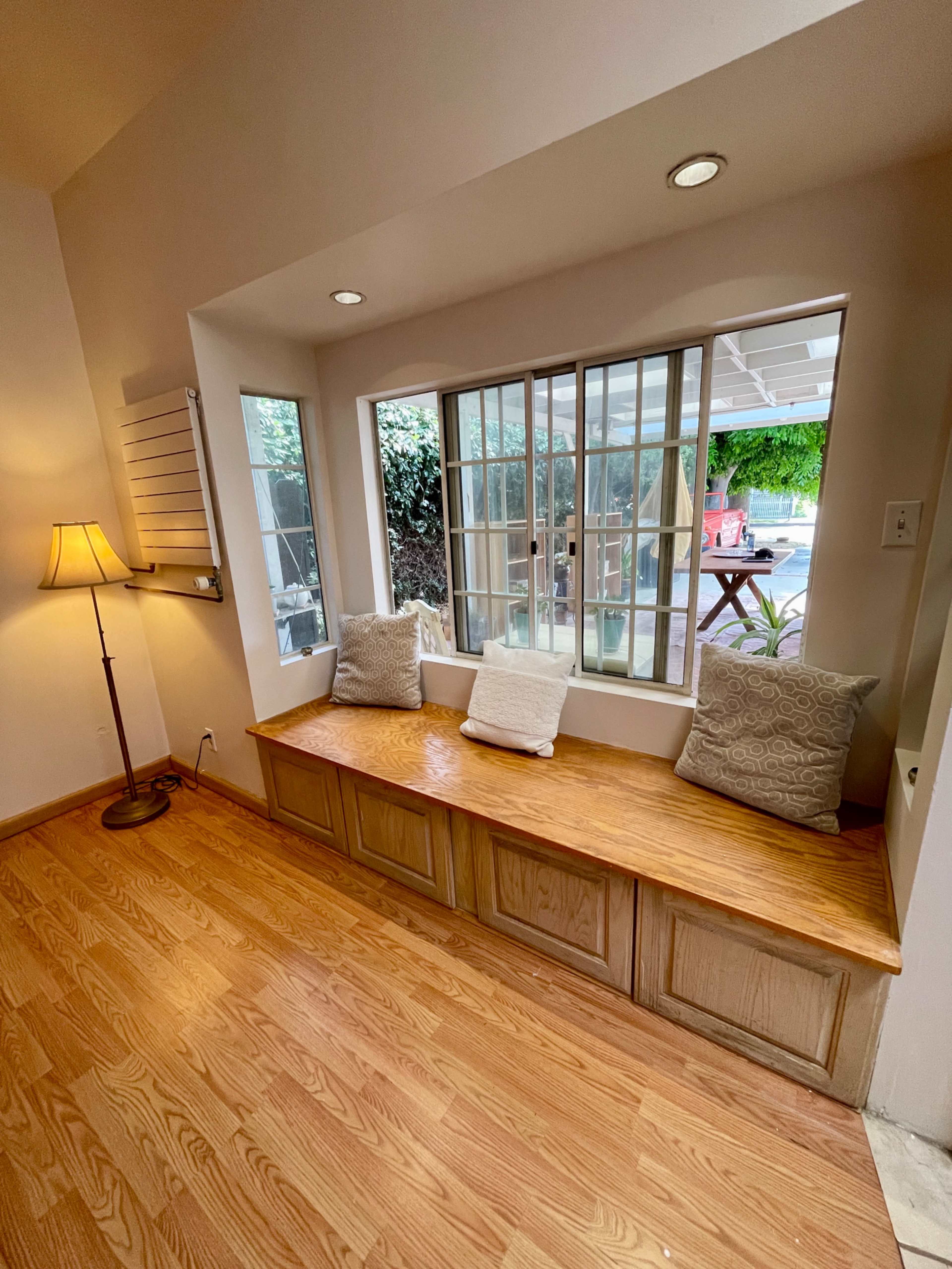 The image shows a cozy window seat area with wooden benches and two decorative pillows, illuminated by natural light from the windows.