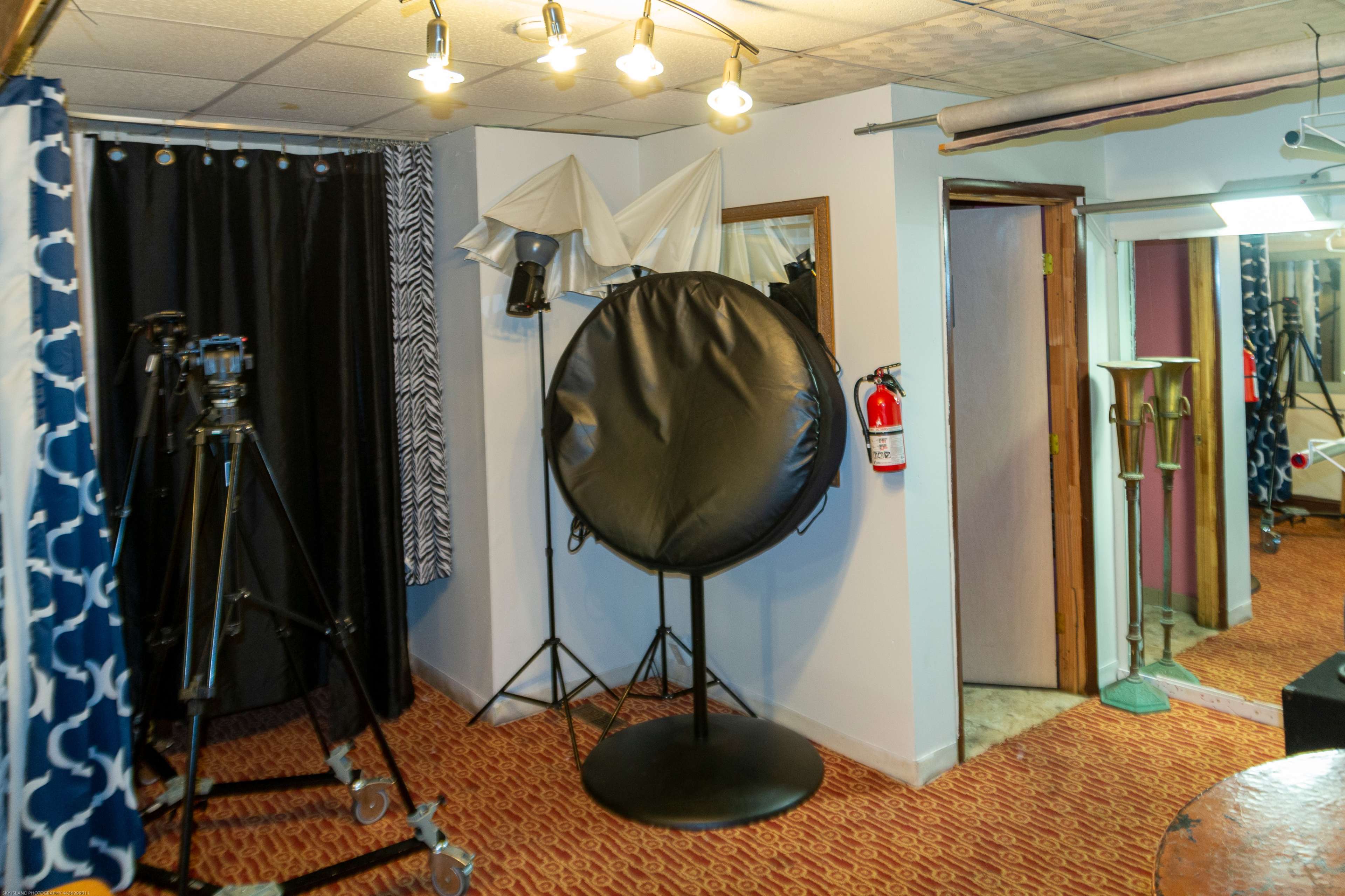 A studio space with photography equipment, including tripods, a reflector, and backdrops, along with an emergency fire extinguisher mounted on the wall.