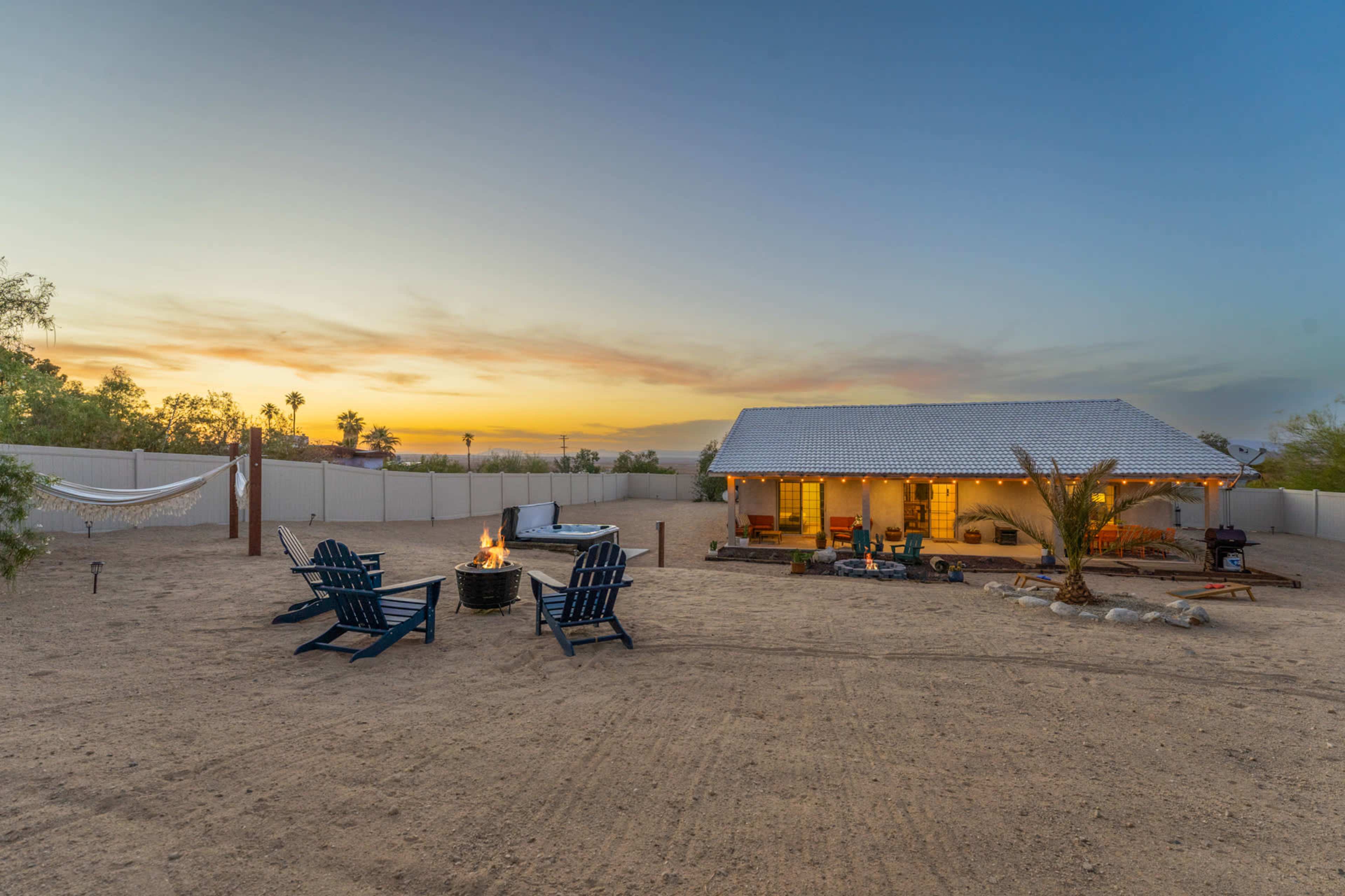 A house with a gray roof is situated in a sandy yard, featuring two blue Adirondack chairs, a fire pit, and a hammock, against a sunset sky.