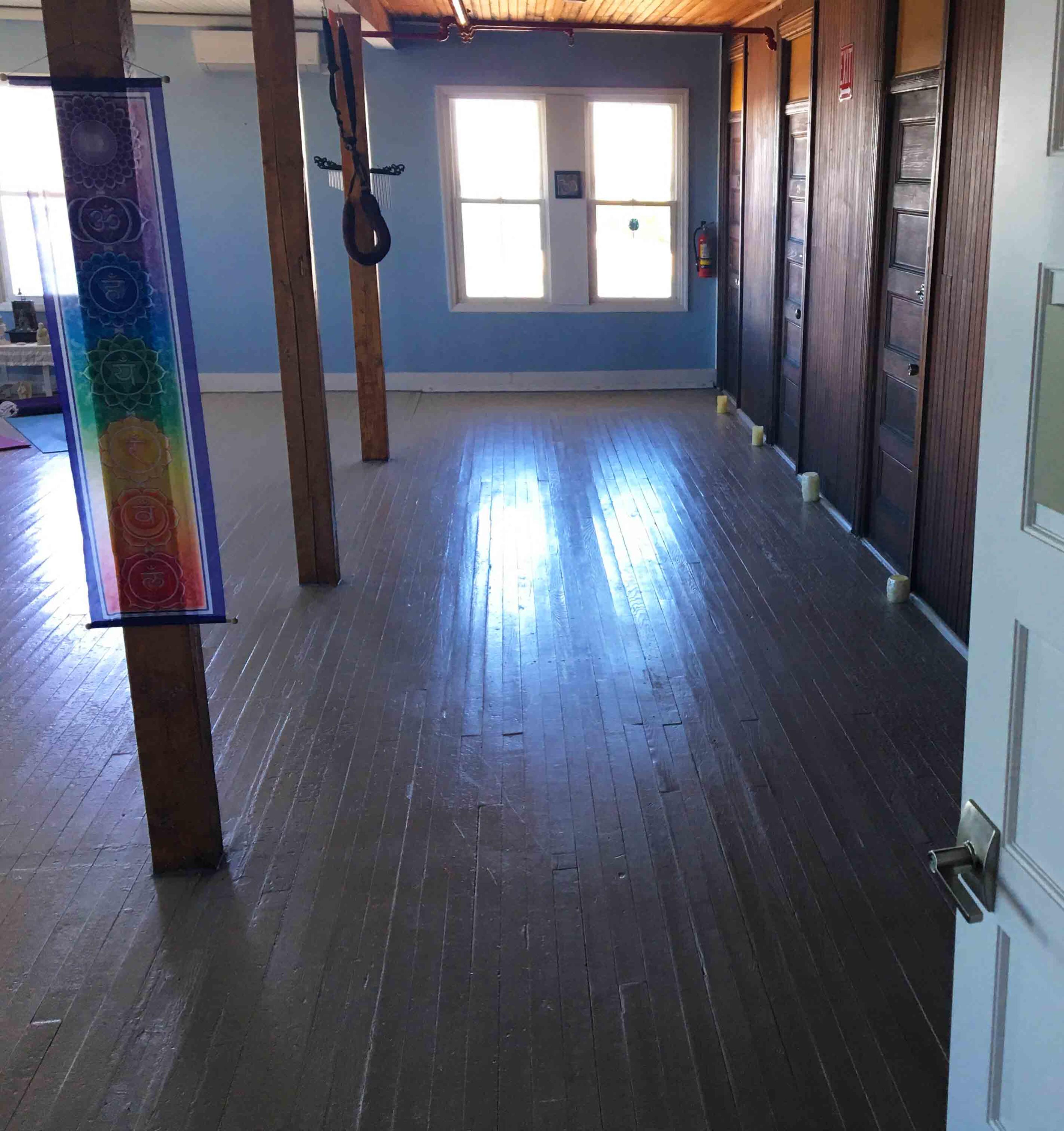 The image shows a narrow hallway with wooden floors, leading to windows on one end and several closed doors on the other.