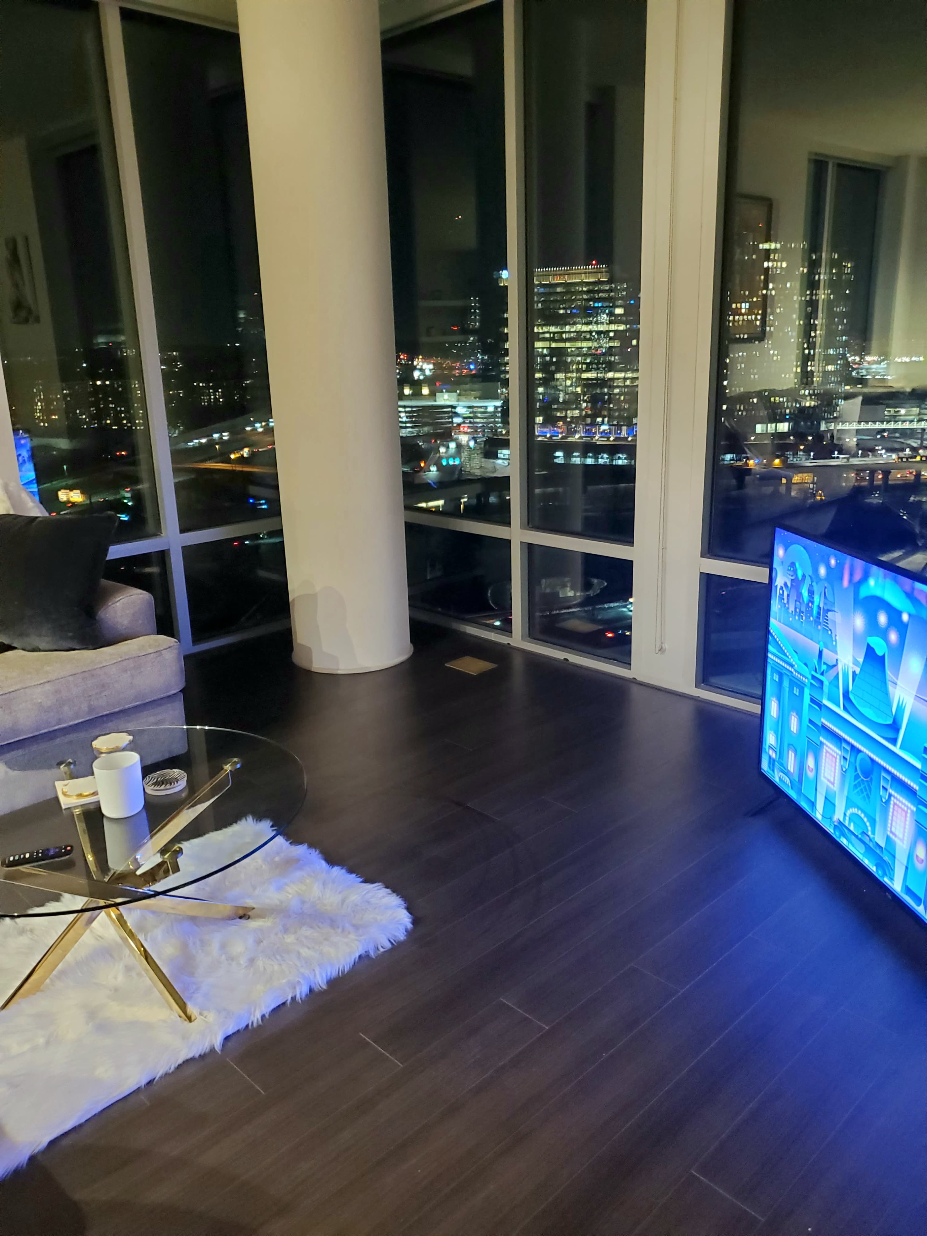 A modern living room with a glass coffee table, a sofa, and a large TV, overlooking a city skyline at night.