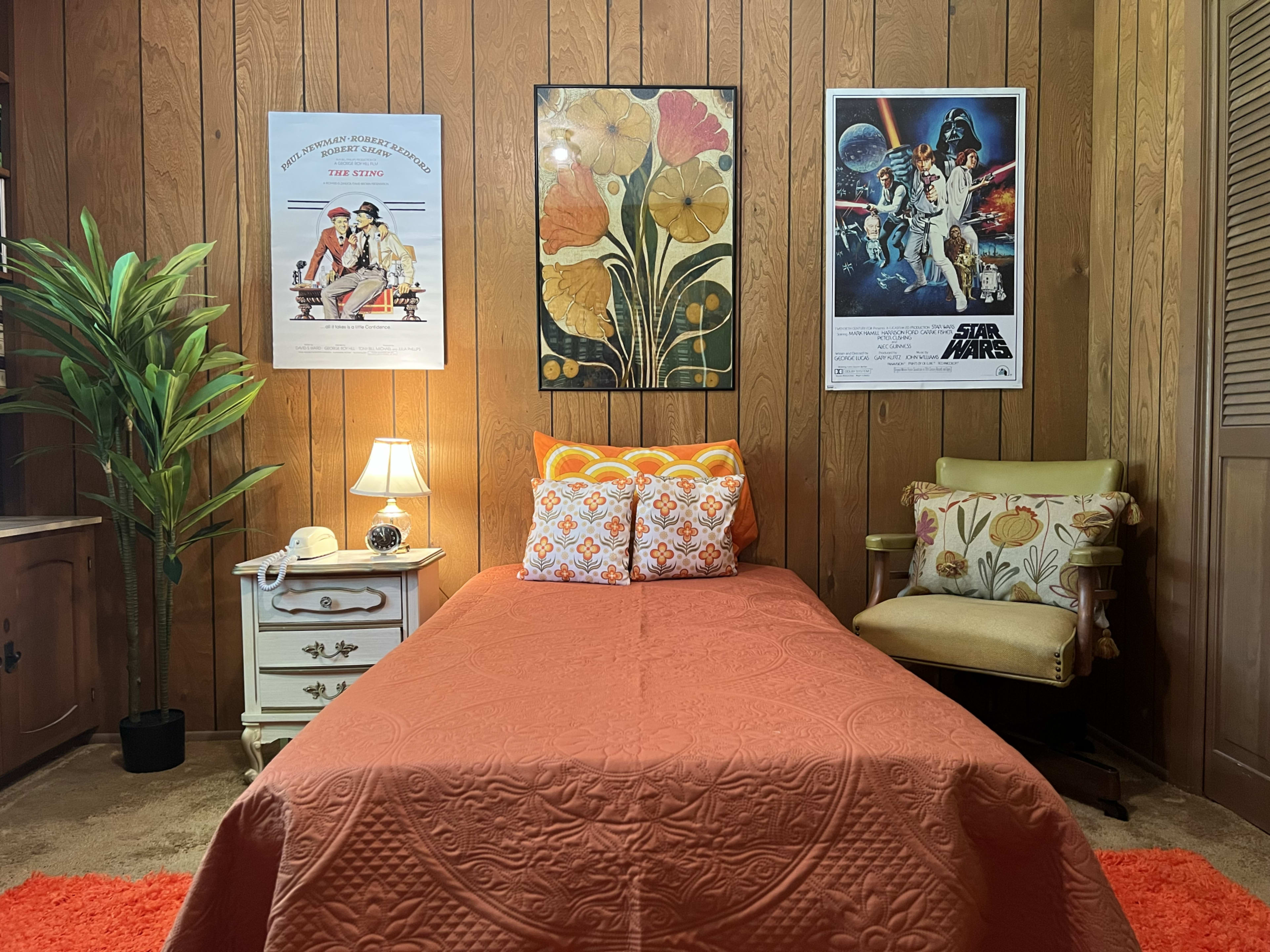 The room features a bed with an orange quilt, surrounded by wooden paneling, a small lamp, plants, and framed posters of floral designs and Star Wars.