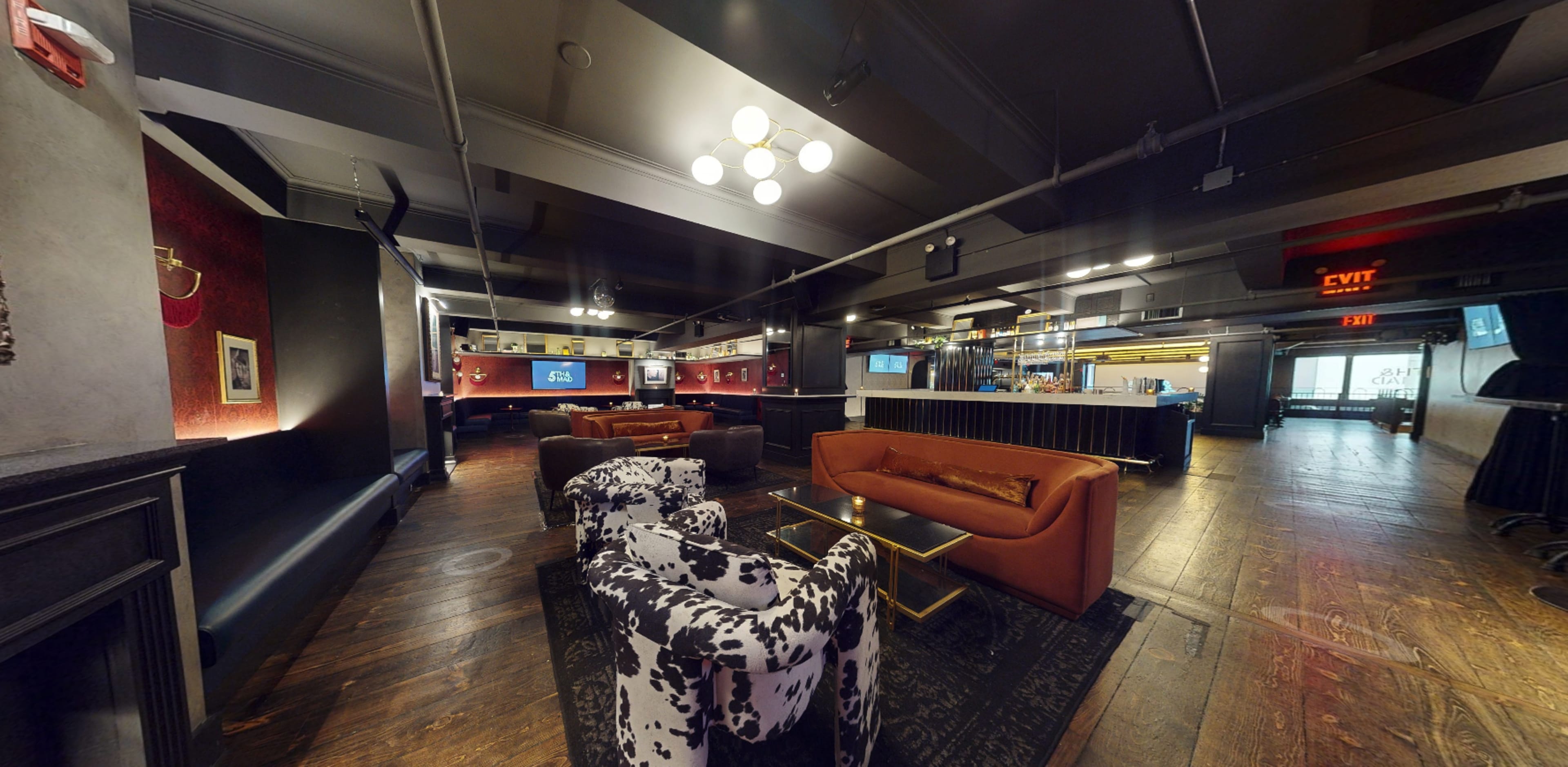 The image shows a modern lounge area featuring various seating options, including a brown couch and patterned chairs, with dim lighting and a bar in the background.