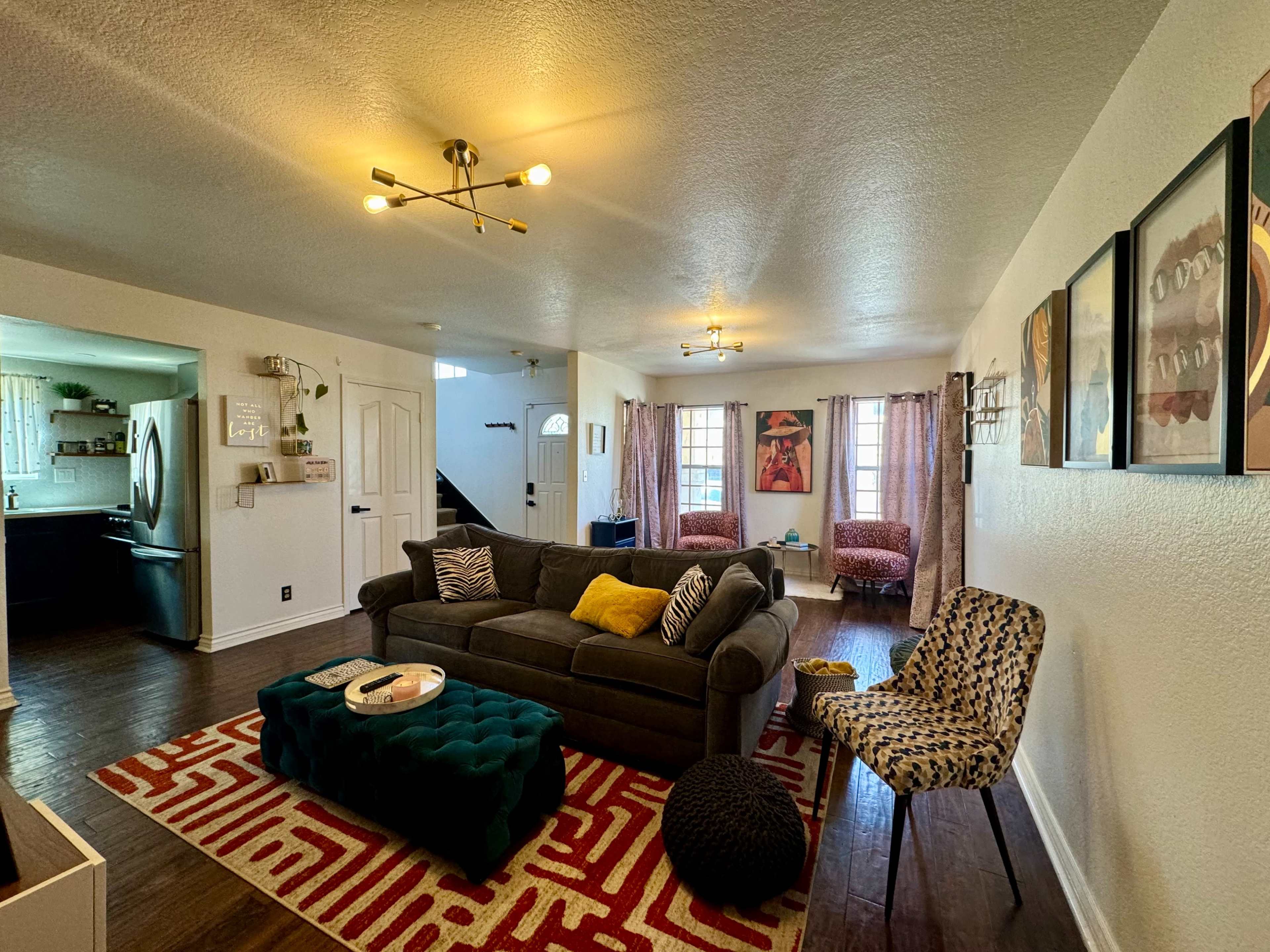 A living room features a gray sofa, colorful throw pillows, a tufted green ottoman, and patterned chairs, with artwork on the walls and hardwood flooring.