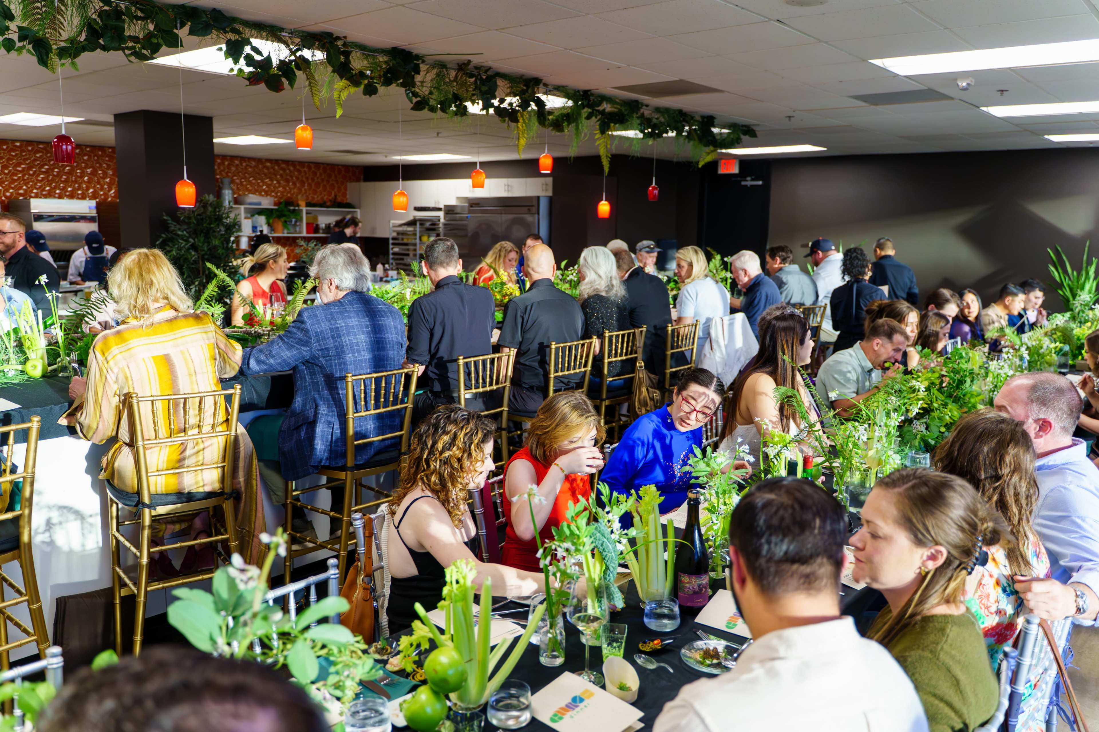 A large group of people attends a dinner event in a decorated banquet space, sitting at a long table adorned with greenery and floral centerpieces.