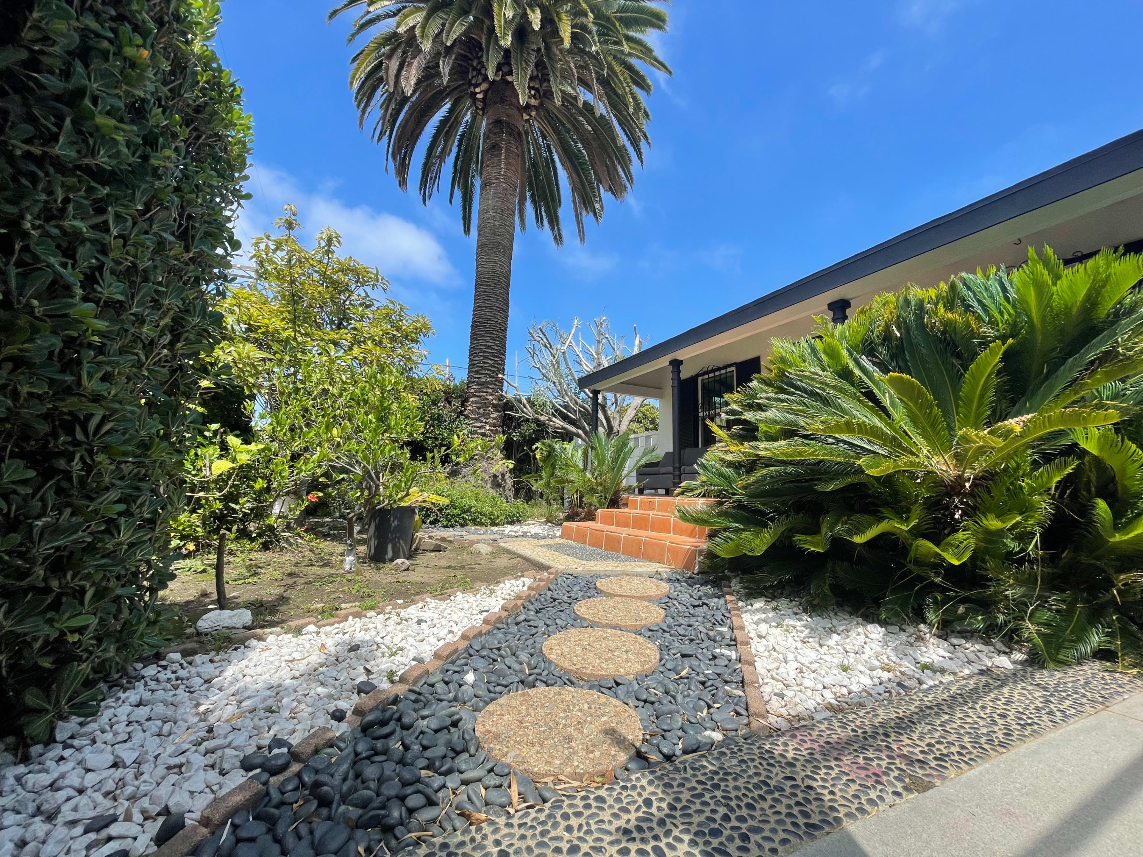 A landscaped pathway made of stones and pebbles, leading towards a house surrounded by palm trees and lush greenery.