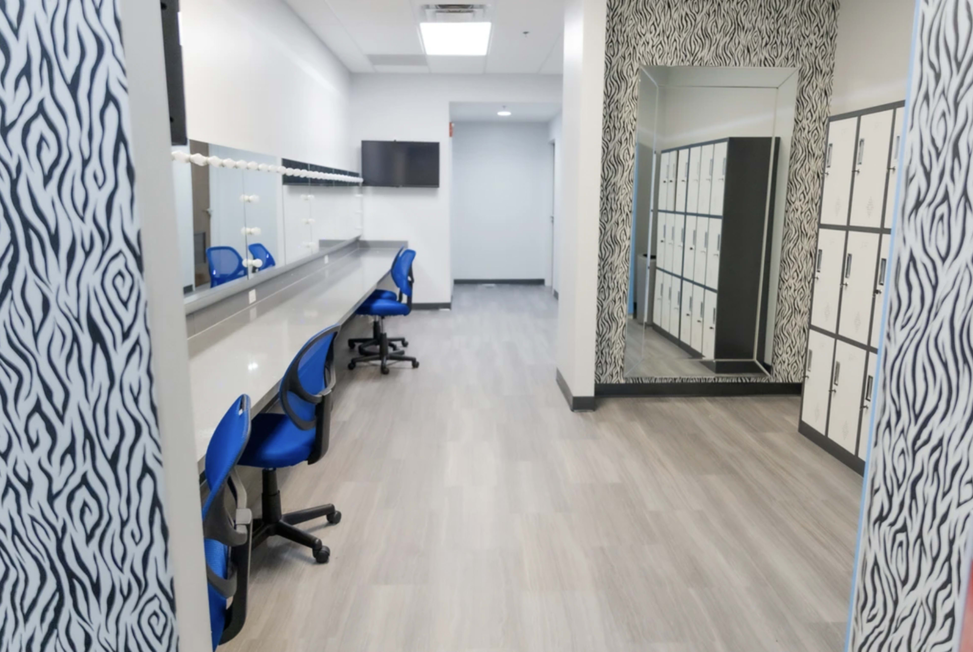 A spacious changing room with blue chairs, a long vanity area with mirrors, a television on the wall, and lockers on one side.