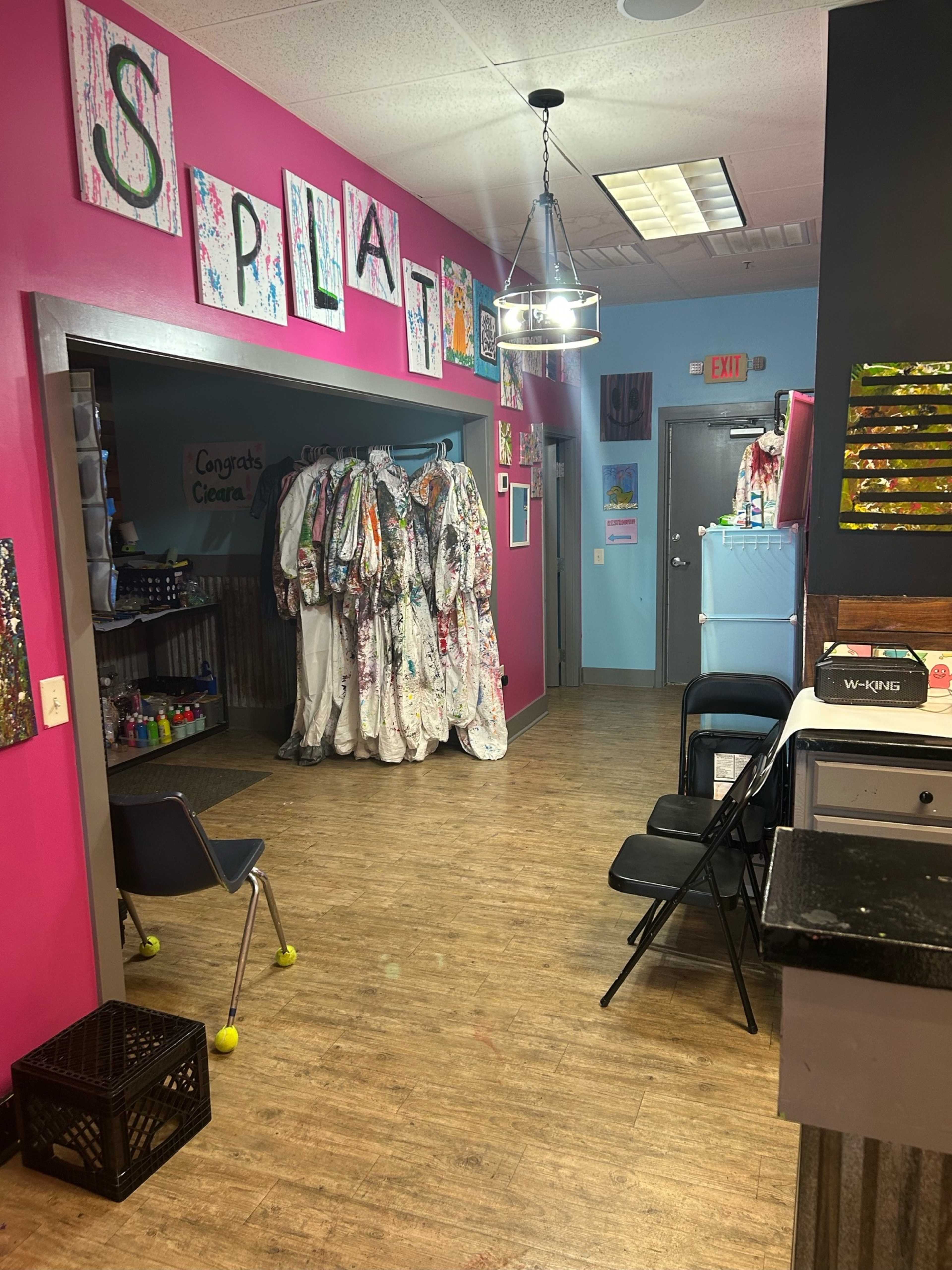 A brightly colored art studio with a corridor leading to a room adorned with splattered paint and hanging artist smocks.