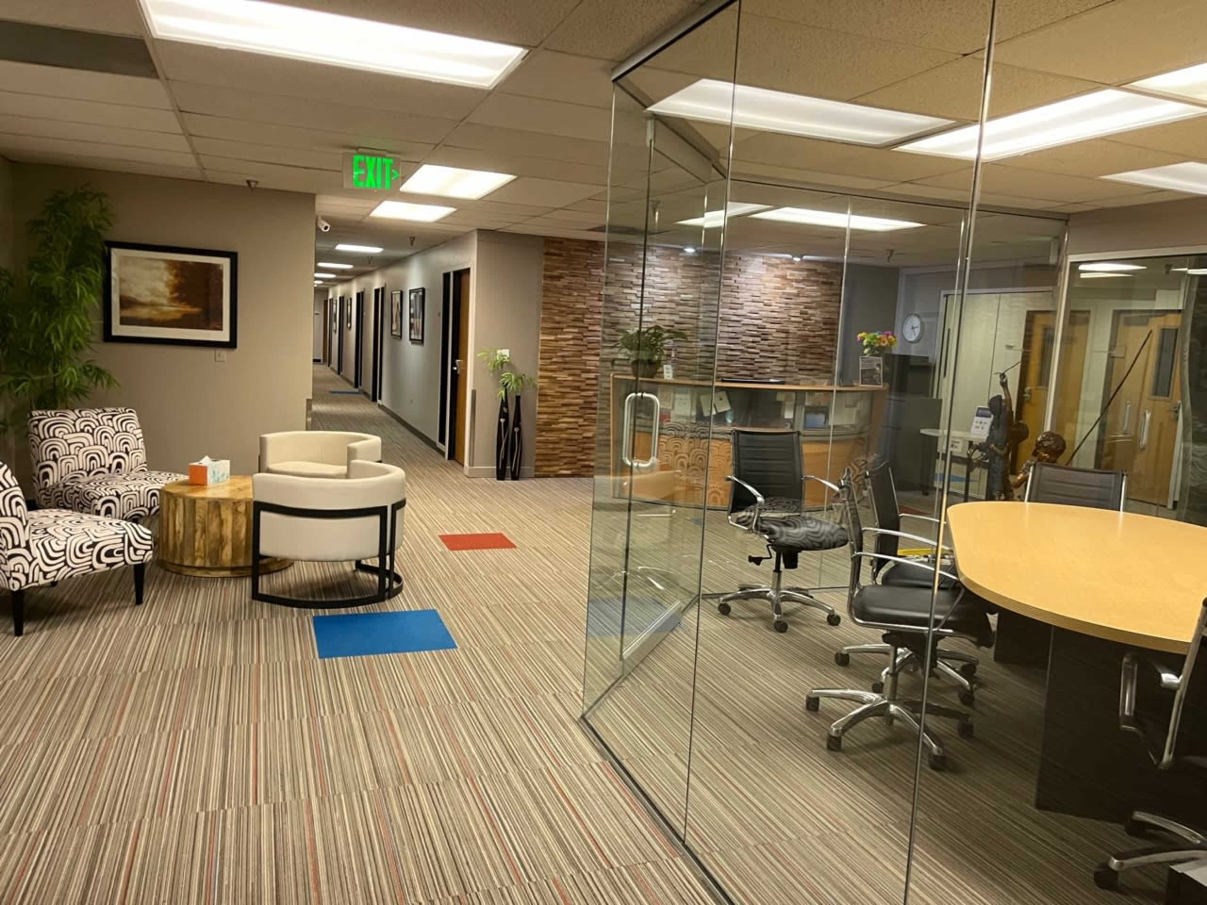 The image shows a modern office space featuring glass walls, a conference table, chairs, and a waiting area with patterned and solid color seating.