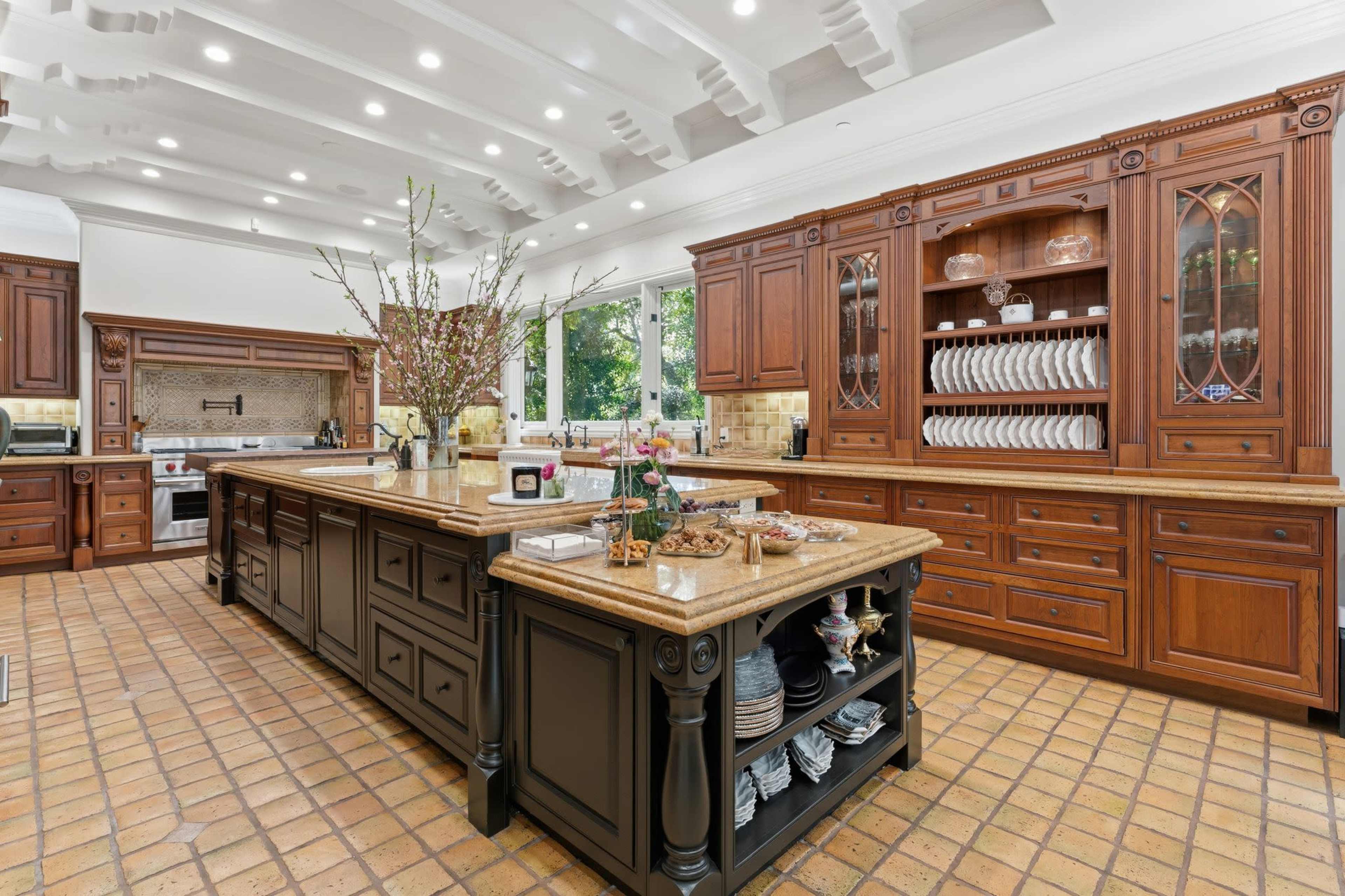 The image shows a large kitchen featuring dark wooden cabinetry, an expansive central island, and terracotta tile flooring.