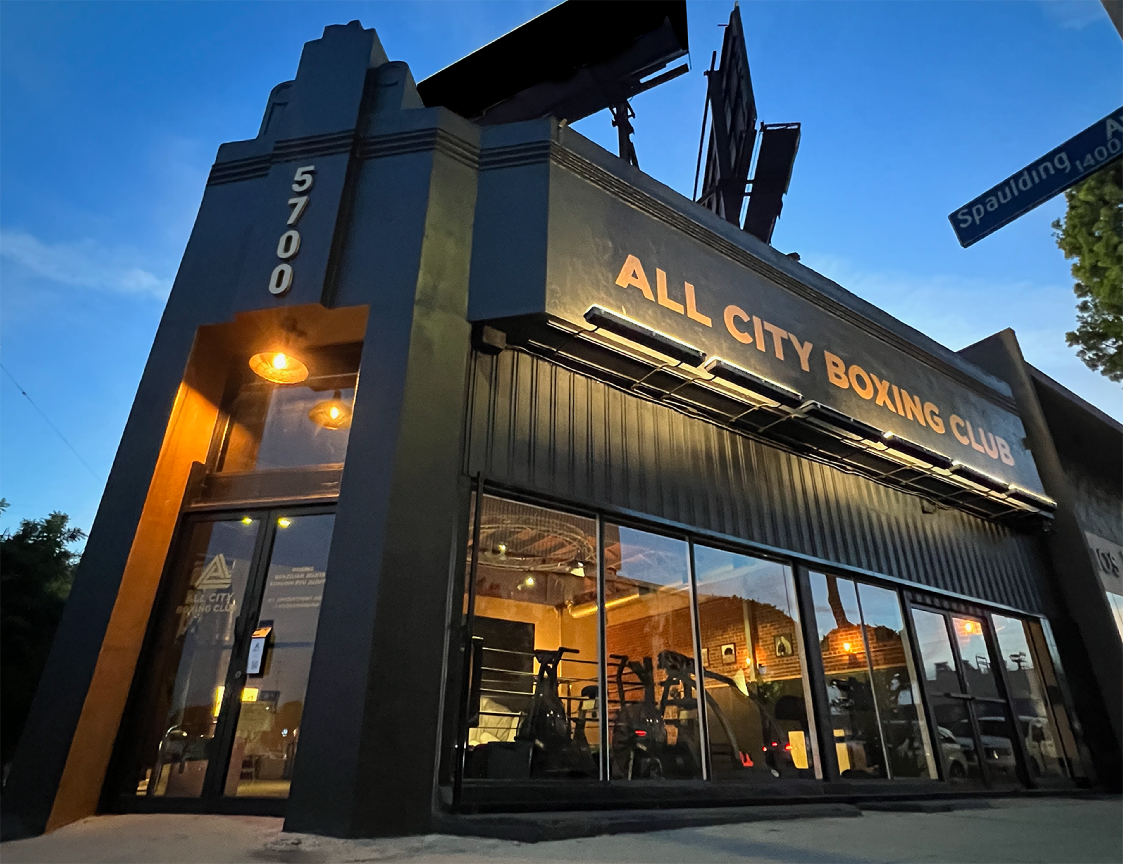 The exterior of the All City Boxing Club, featuring large windows and a sign, set against a twilight sky.