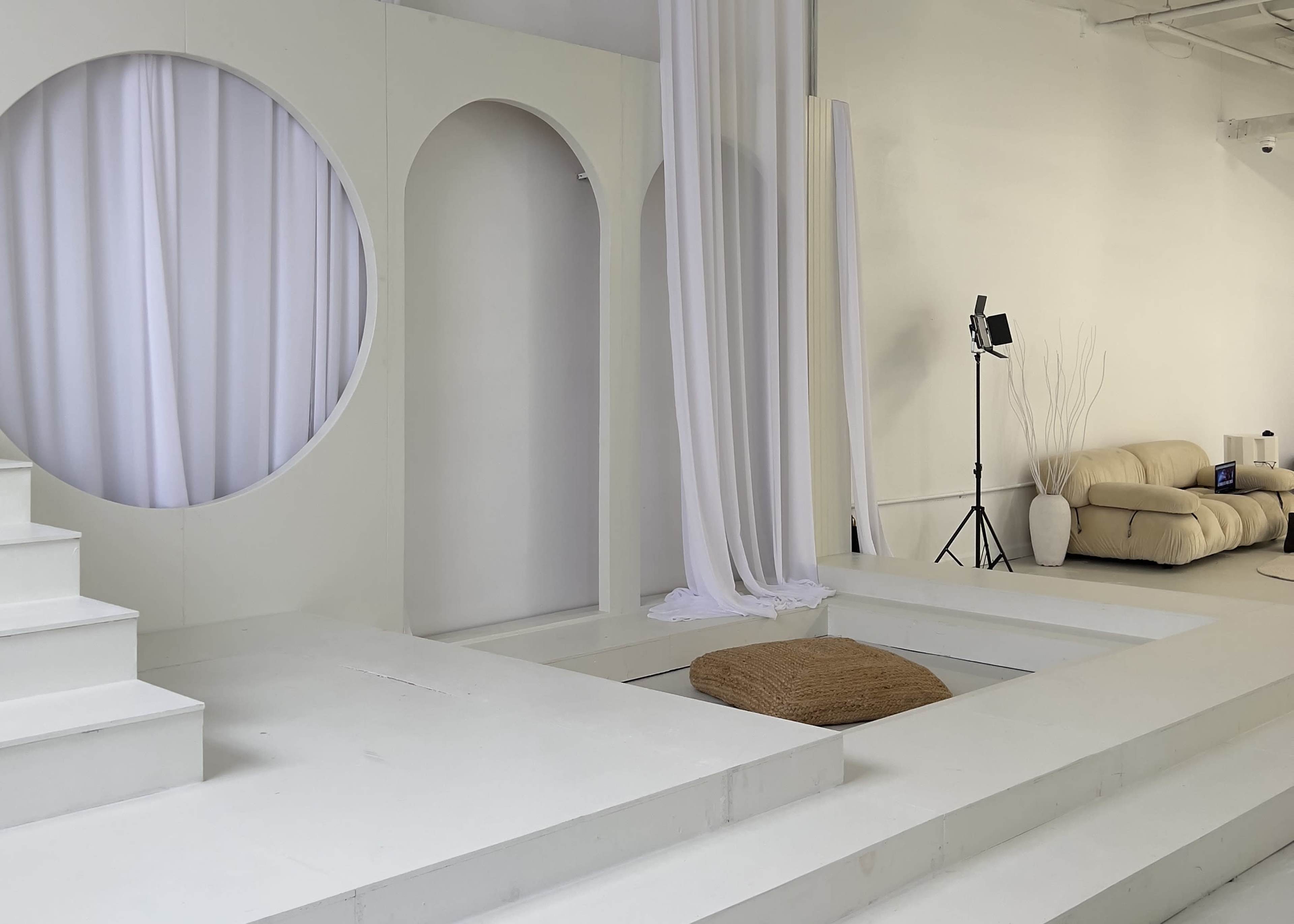 A minimalist interior space with white walls, a circular window, steps leading to a sunken area with a circular mat, and a beige sofa in the corner.
