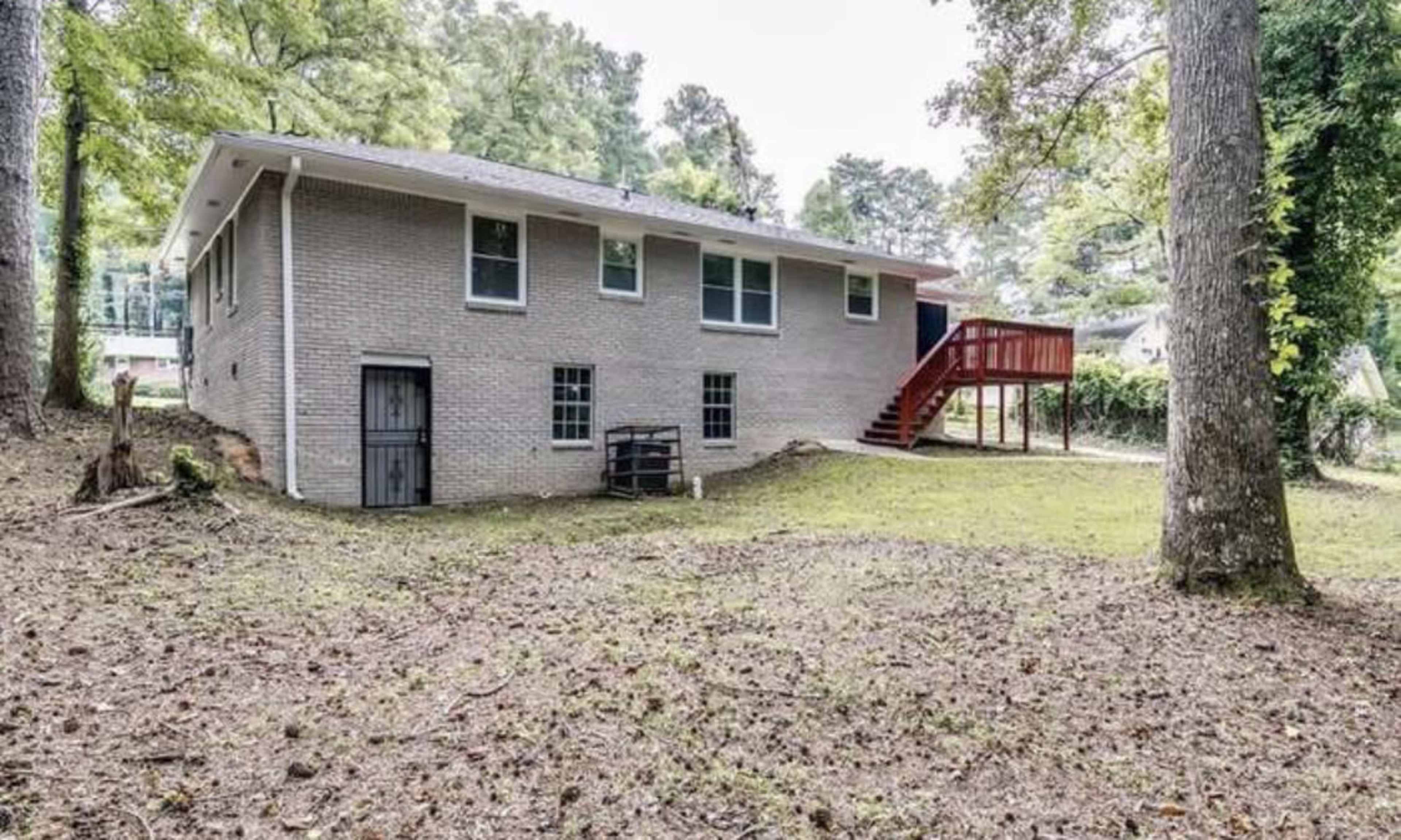The back of a brick house with a wooden deck and stairs, surrounded by a grassy yard and trees.