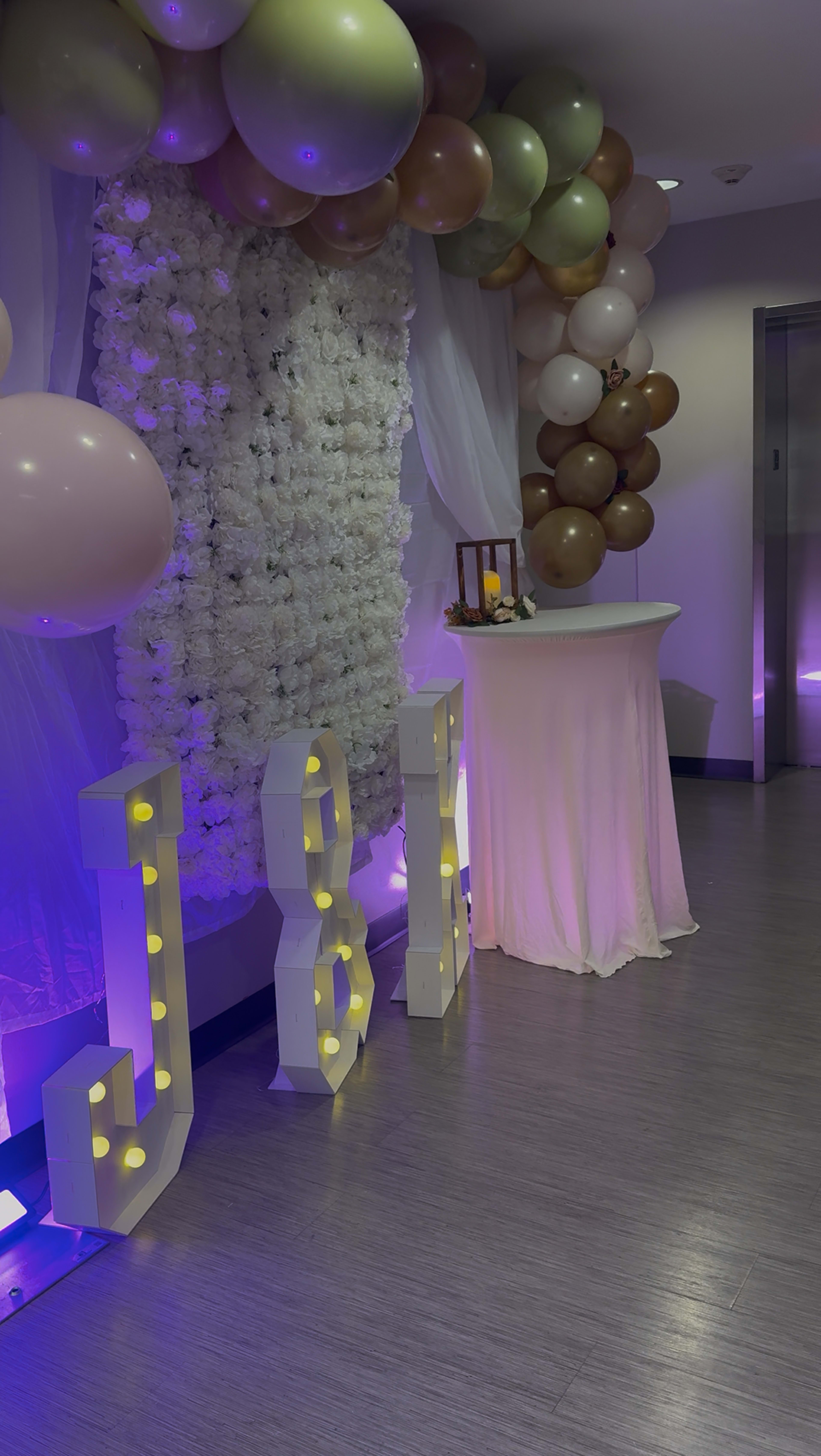 The image shows a decorated event space featuring a wall of artificial flowers, a table with a light-colored tablecloth, and illuminated letters "J&J" beside a balloon display.