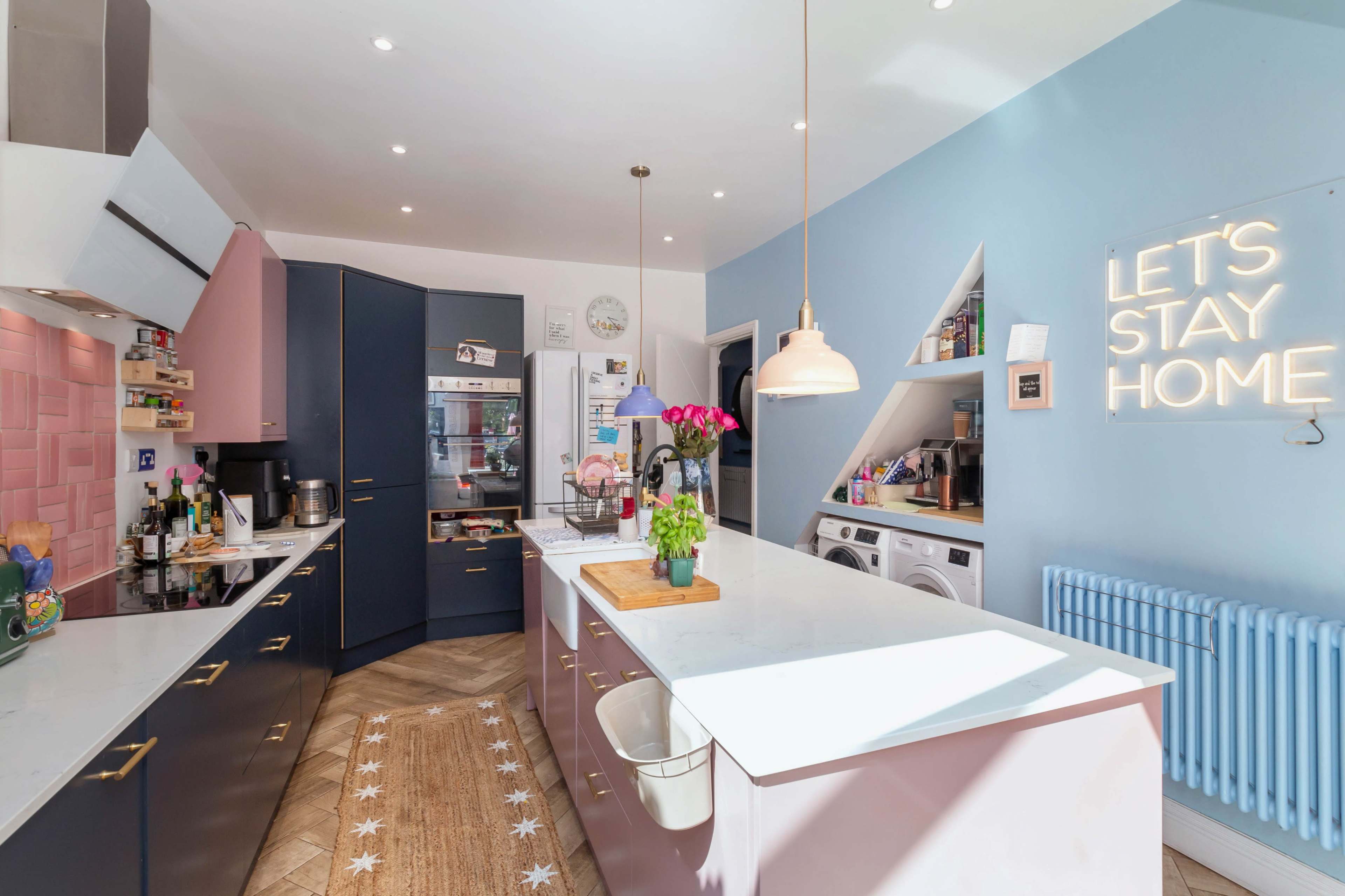 A modern kitchen features a combination of navy blue and pink cabinets, a central island with a cutting board, and a wall with a light-up sign that reads "LET'S STAY HOME."
