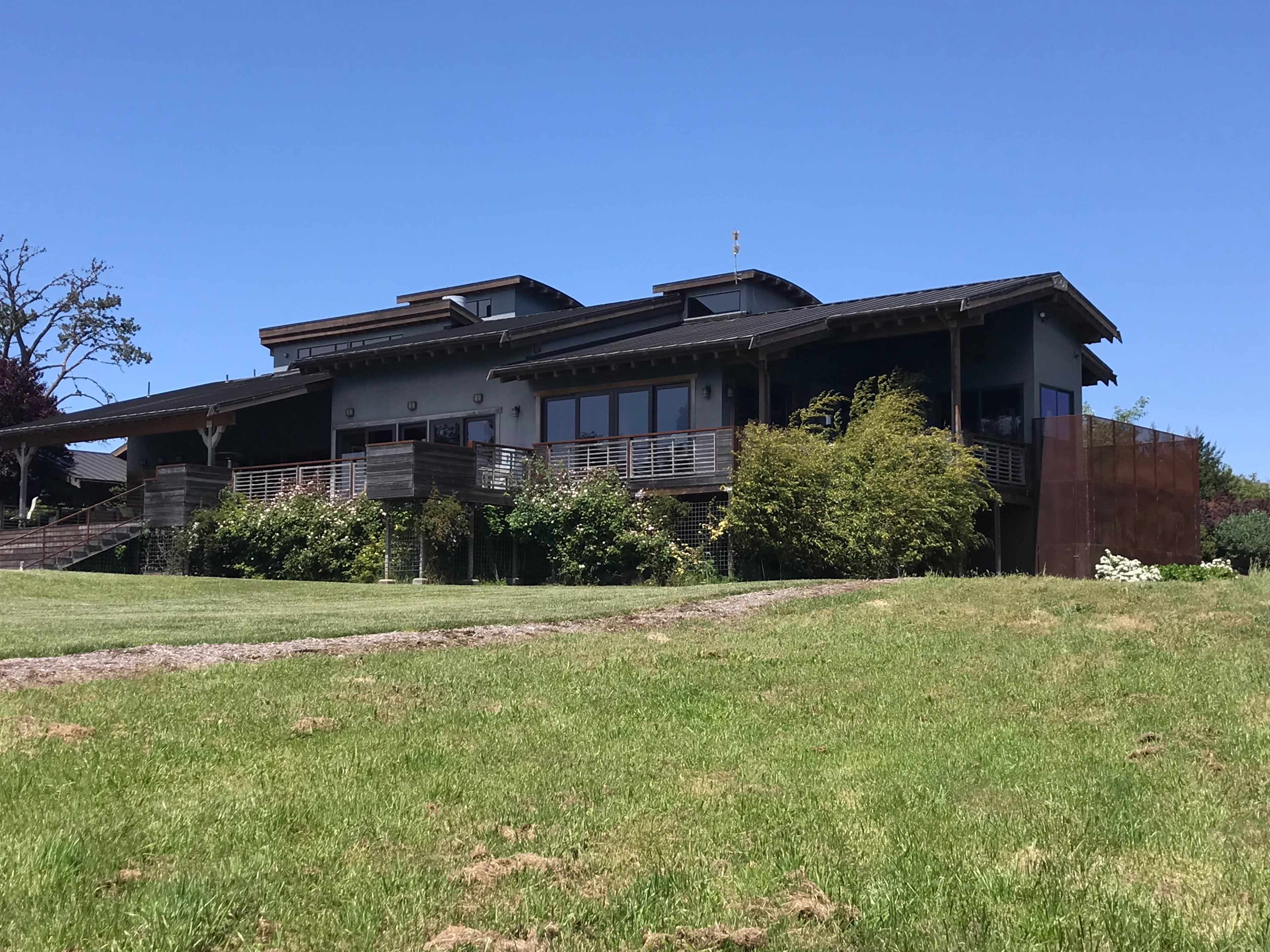 The image shows a large, modern house set on a grassy hillside, featuring a wide deck and multiple stories.