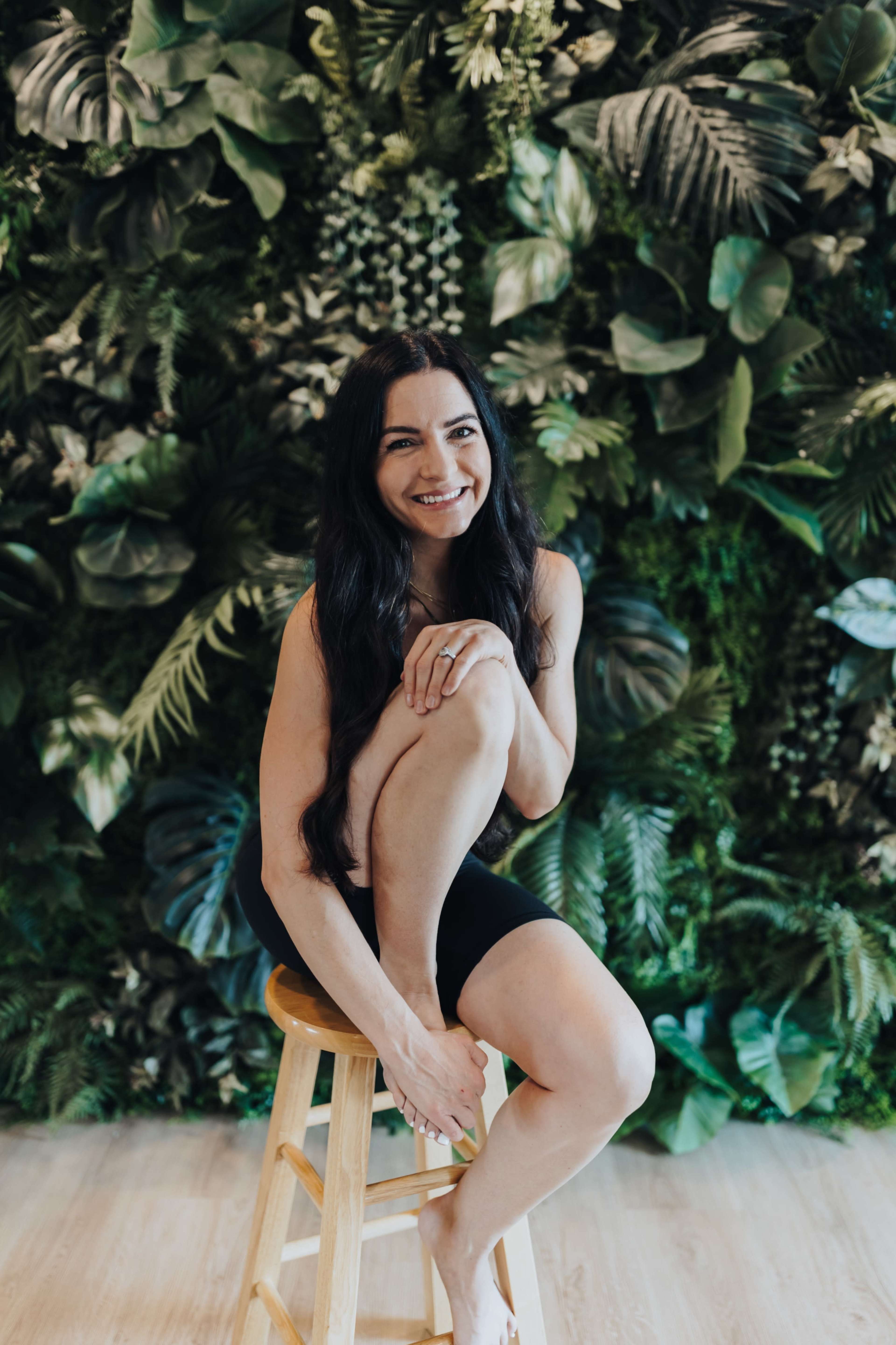 A woman is sitting on a wooden stool against a backdrop of lush green foliage.
