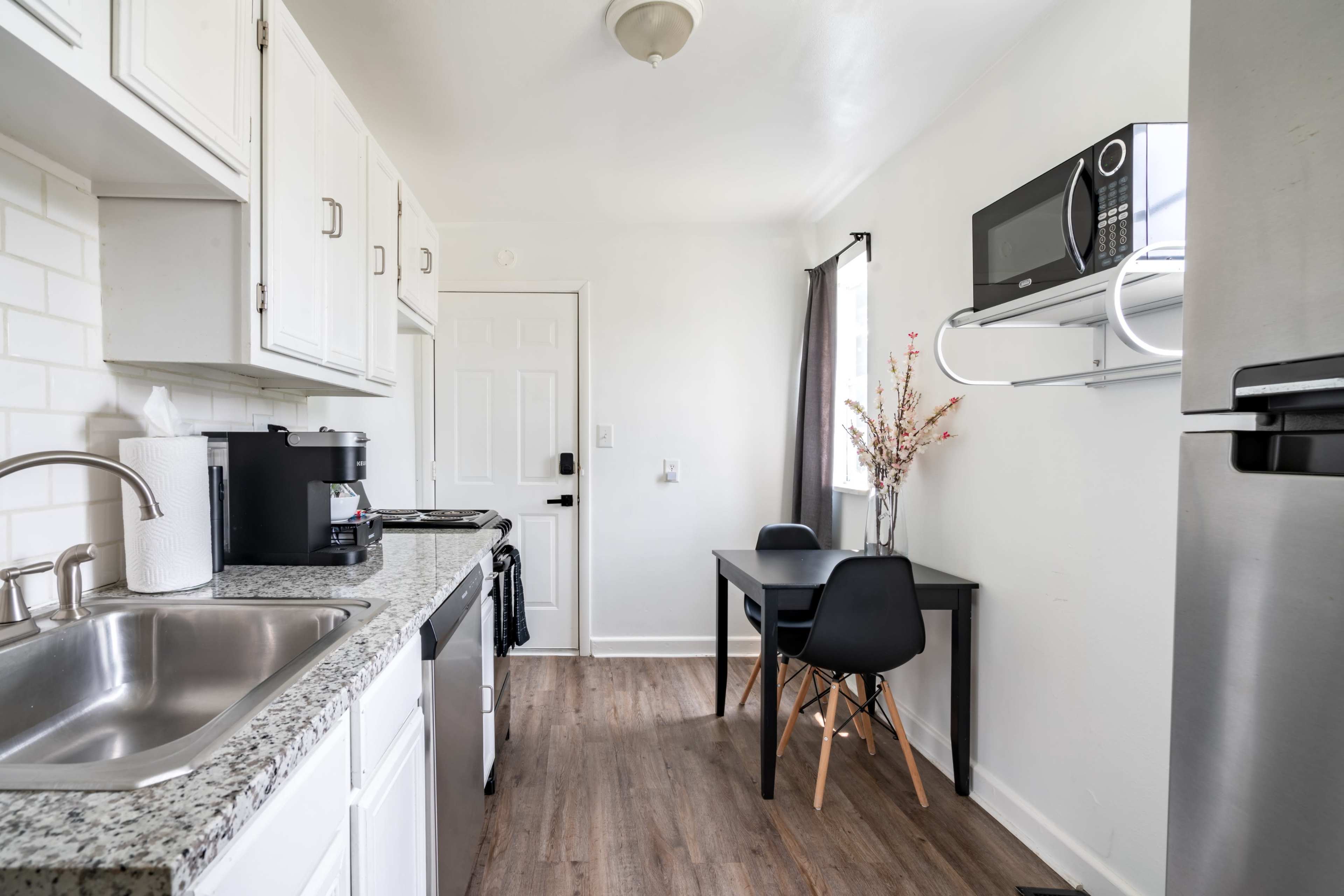 The image shows a compact kitchen with white cabinetry, a stainless steel refrigerator, and a dining area featuring a small table and black chairs.