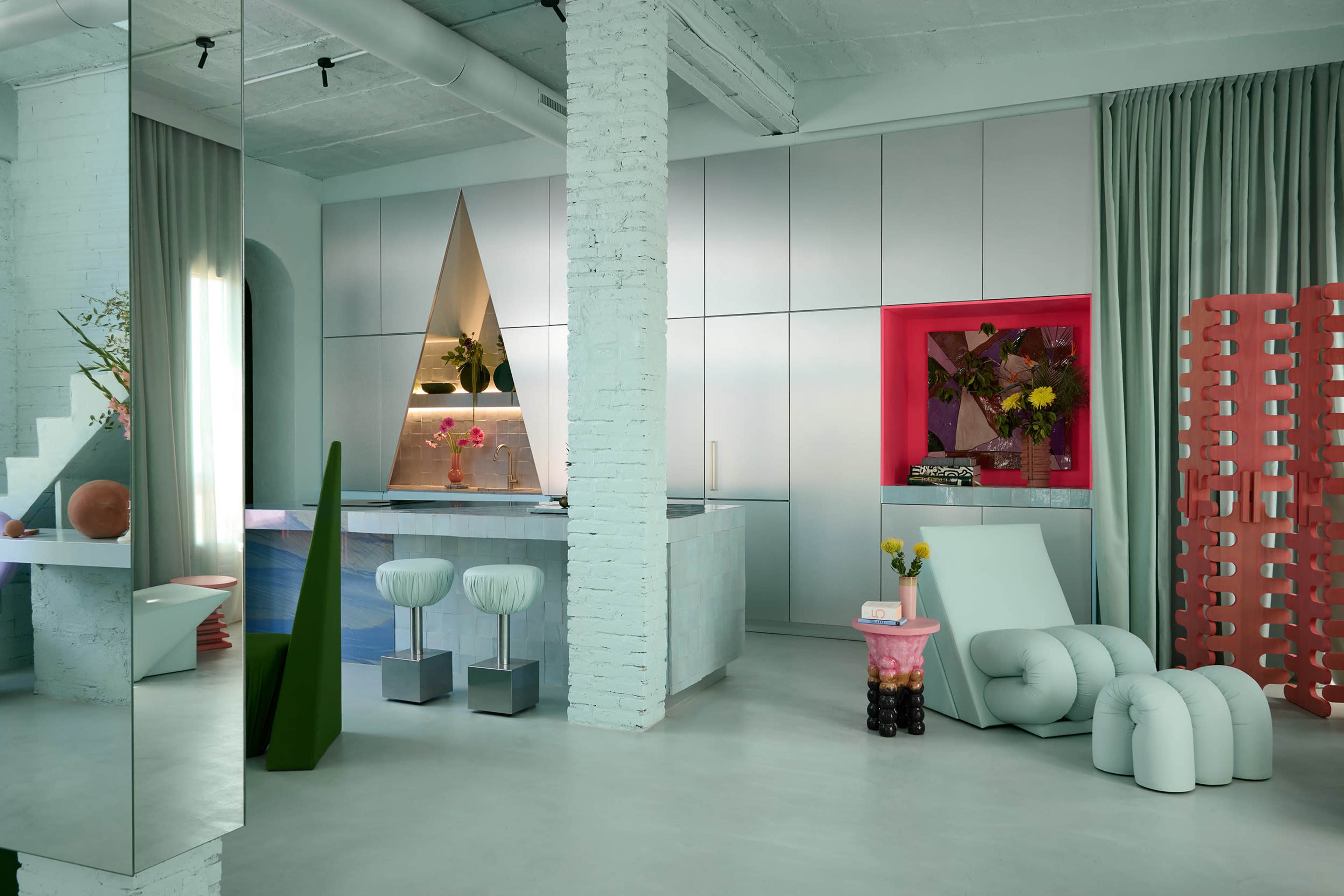 The image shows a modern interior space featuring a kitchen with geometric furnishings, including a triangular accent, colorful decor, and a minimalist design.