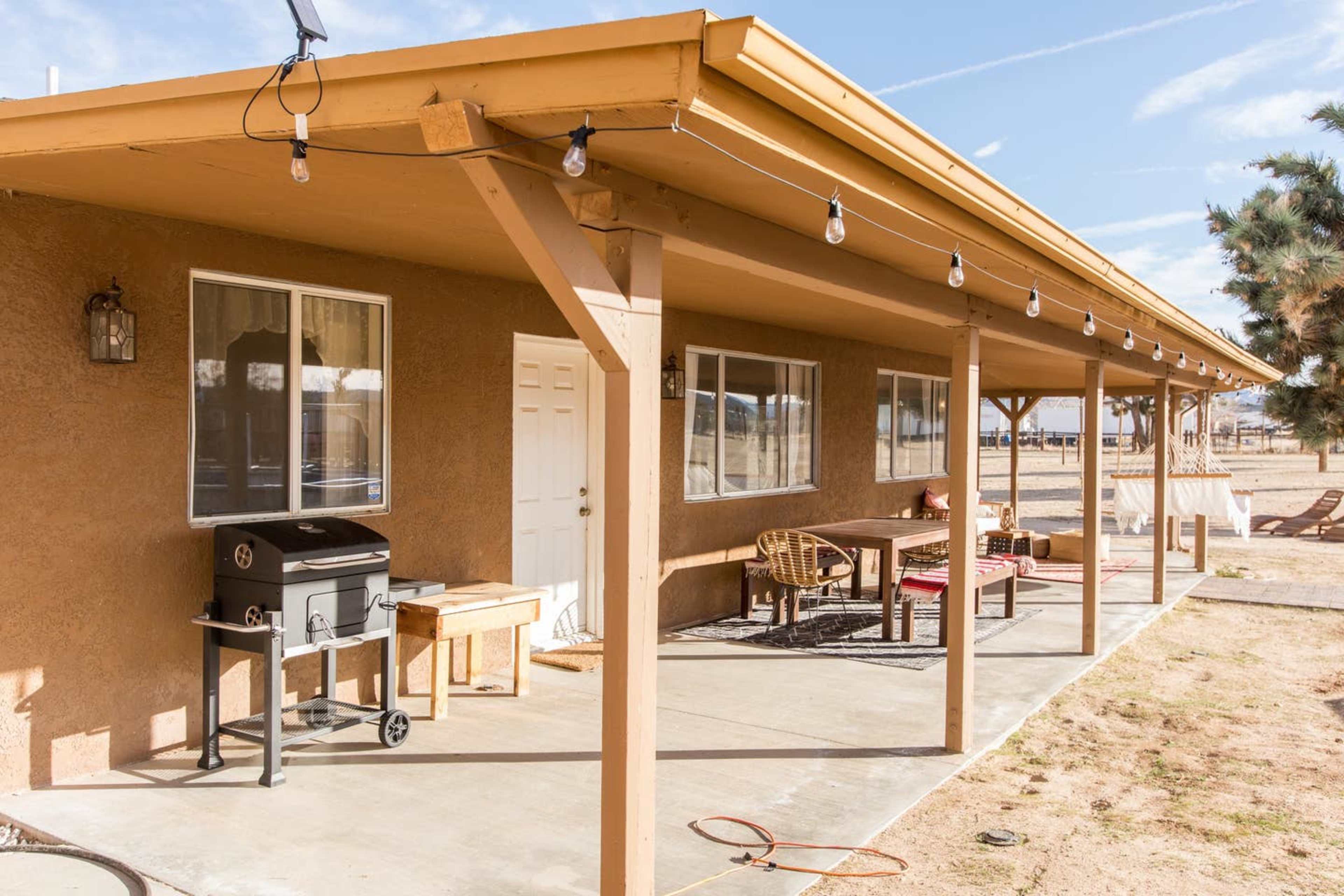 A covered porch with a barbecue grill, a table with chairs, and several windows on a brown stucco building.