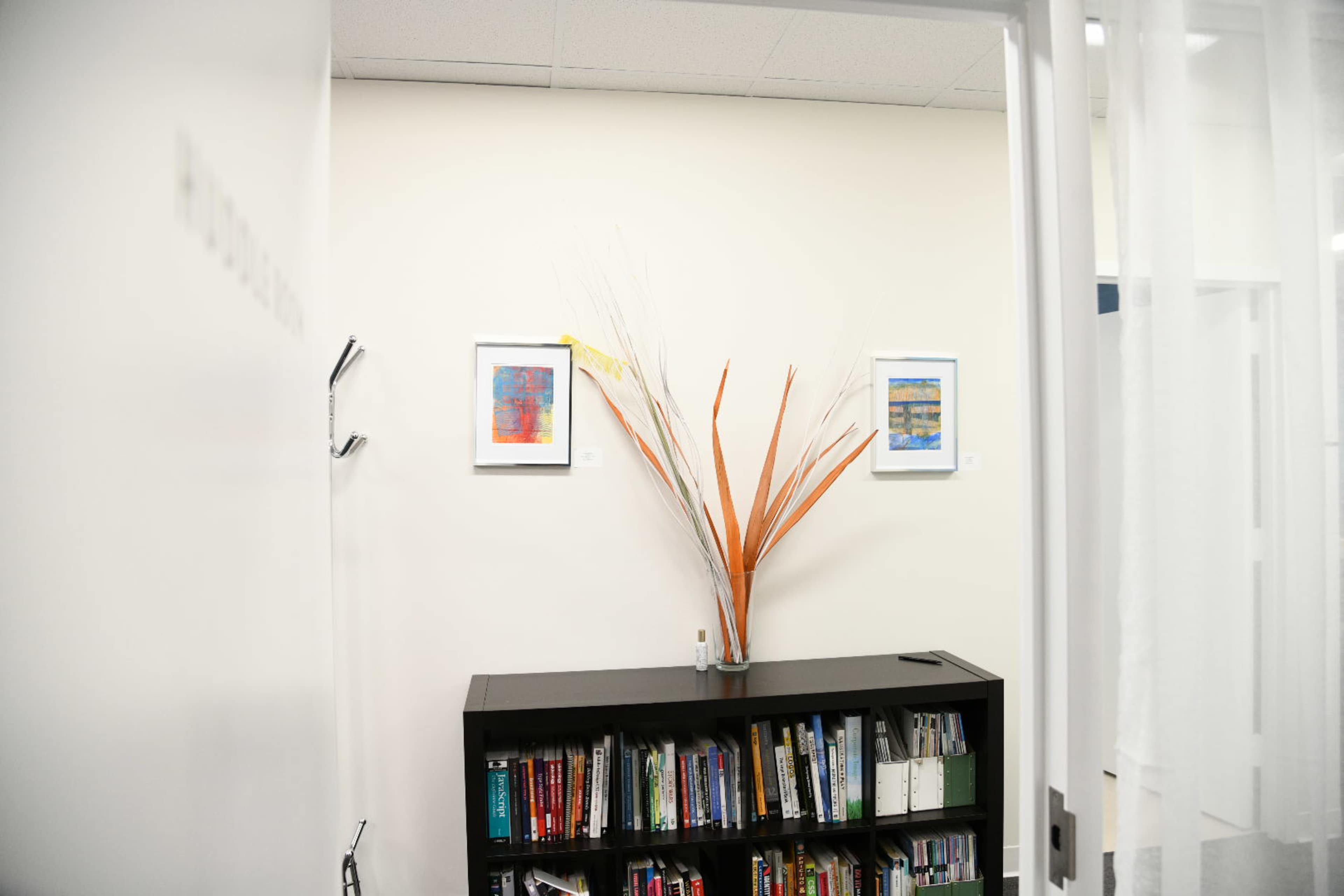 A bookshelf filled with books stands against a wall, featuring framed artwork and a decorative plant arrangement above it.