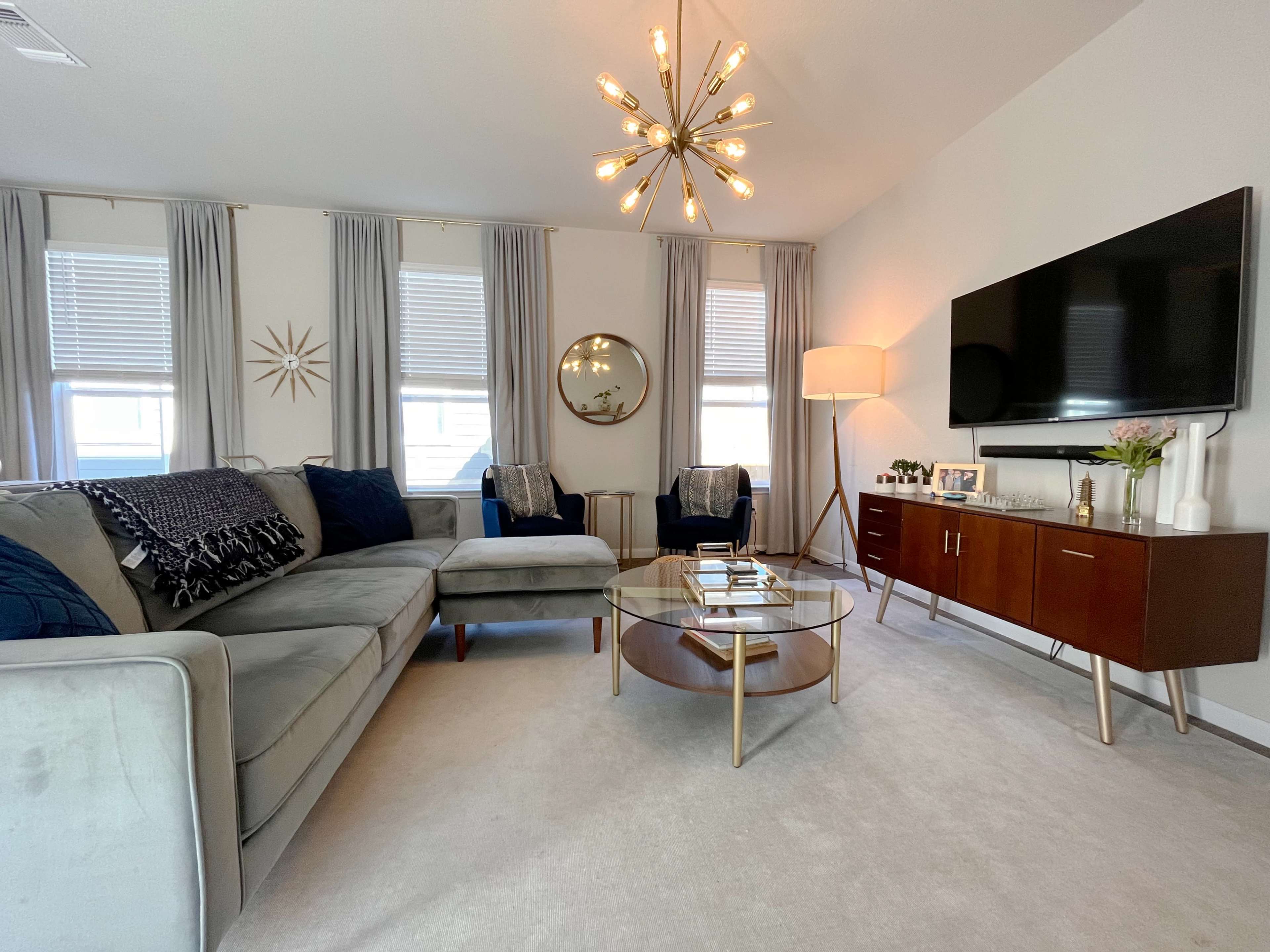 The living room features a gray sectional sofa, a round glass coffee table, and a wall-mounted television, with large windows dressed in light curtains and a mid-century modern light fixture above.