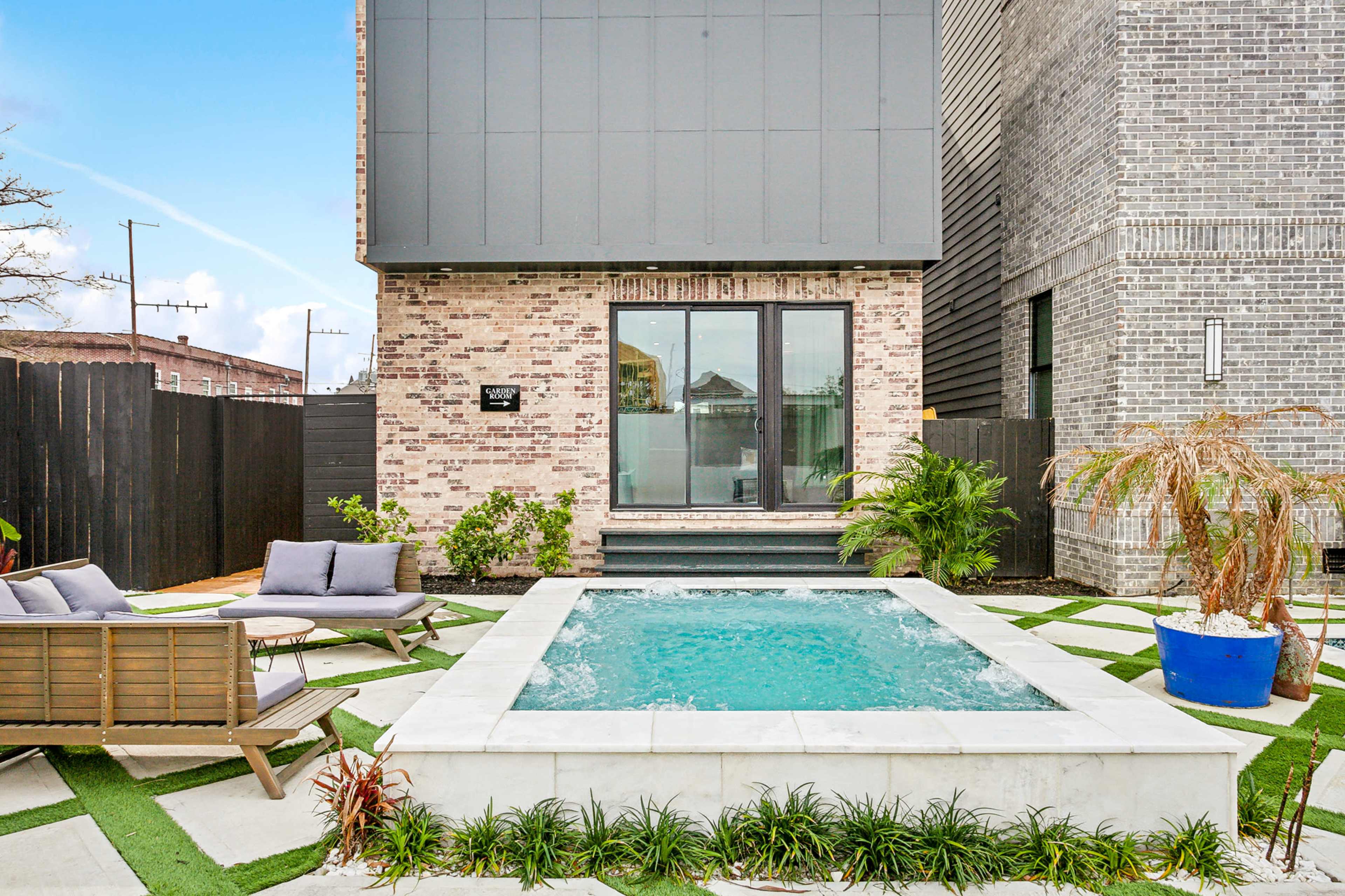 A small, rectangular swimming pool surrounded by stone tiles and seating made of wooden furniture in a landscaped outdoor area.