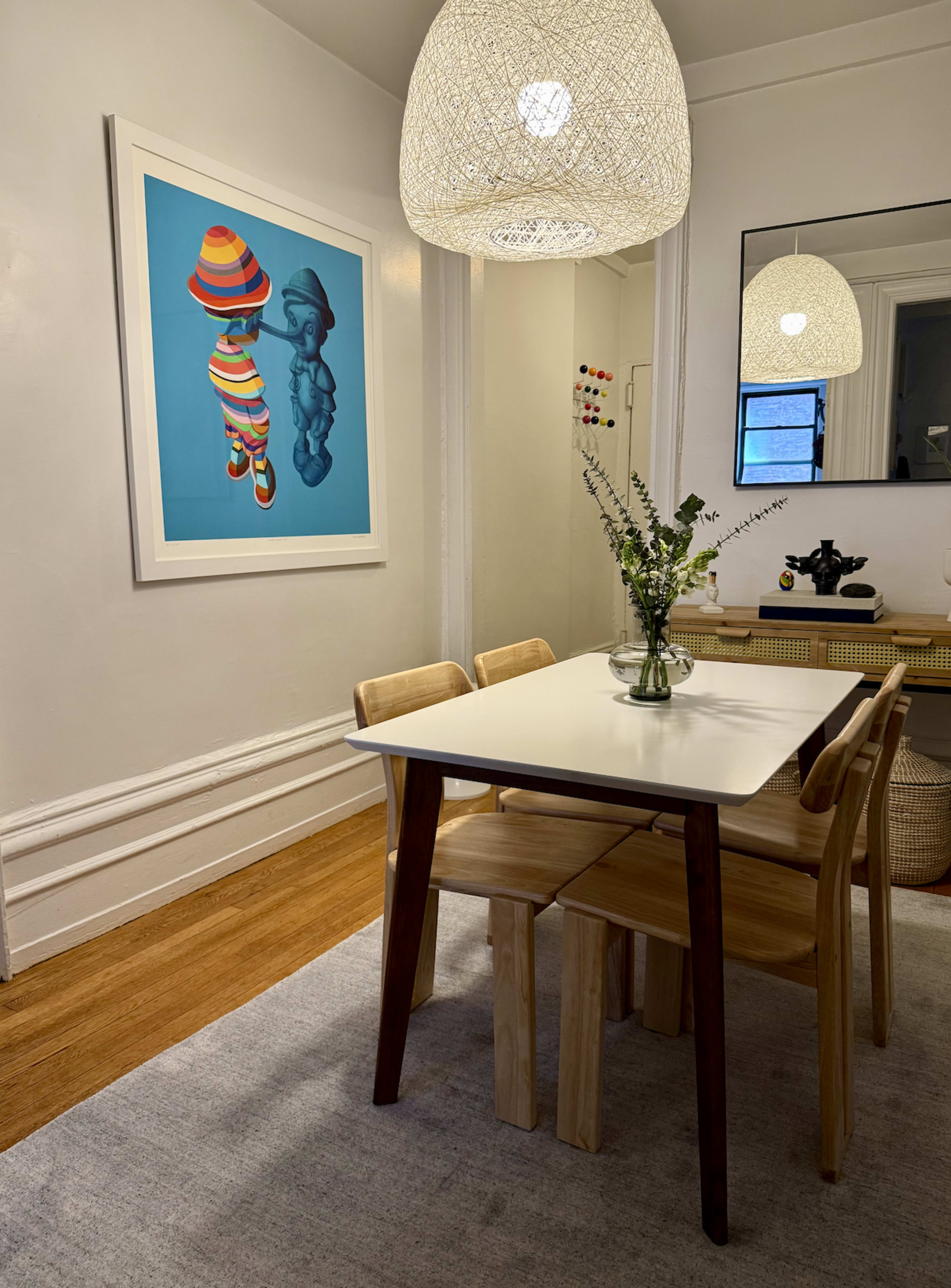 The image shows a dining area with a white table surrounded by wooden chairs, a large colorful artwork on the wall, and a woven light fixture above.
