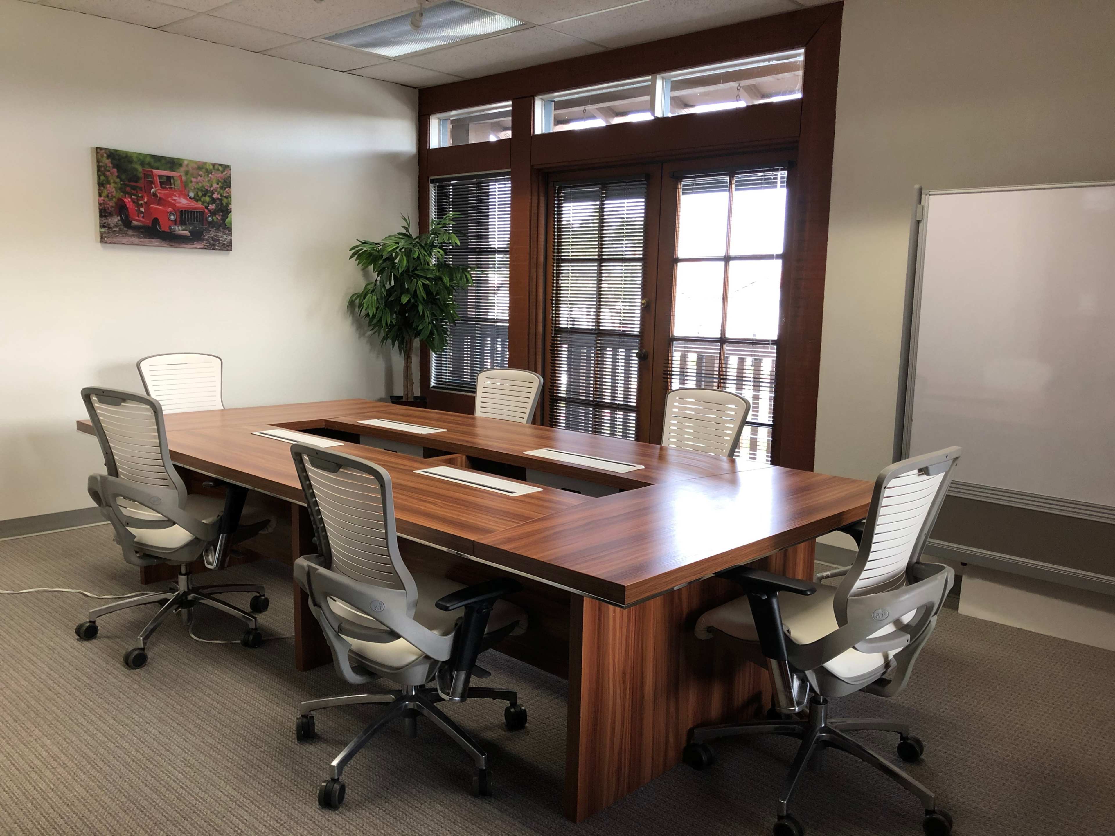 A conference room with a large wooden table surrounded by five ergonomic chairs, and large windows with blinds letting in natural light.