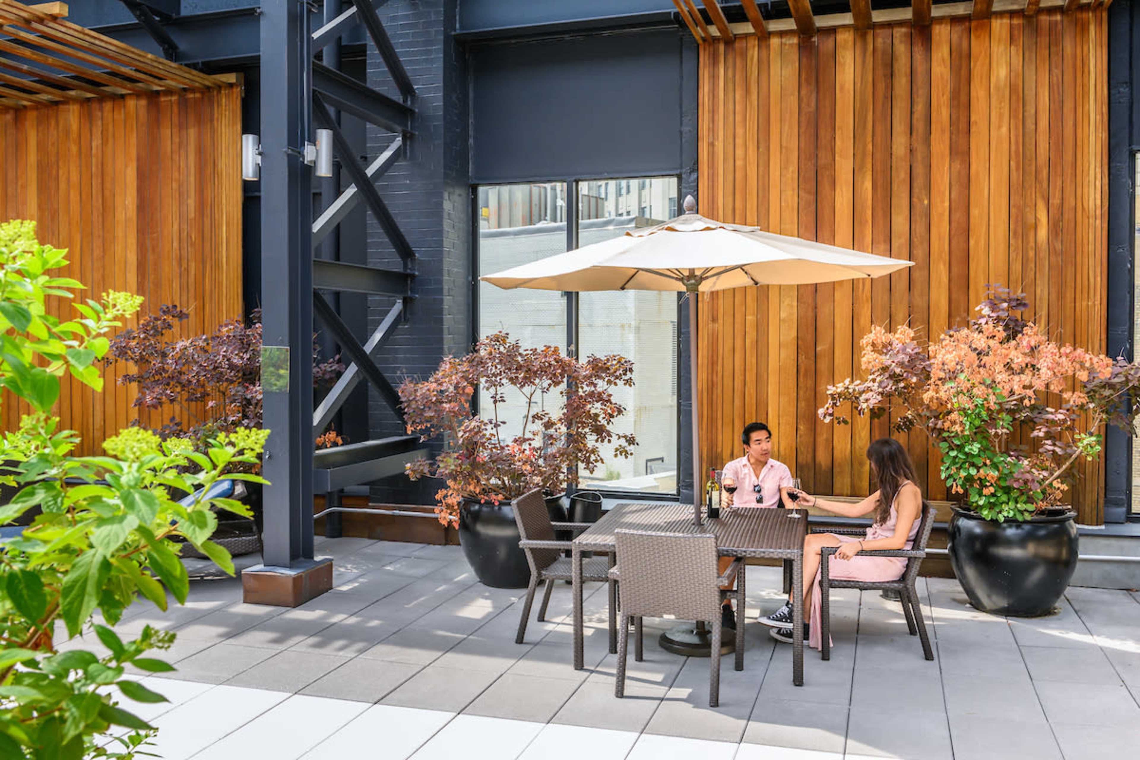 Two people sit at a table under an umbrella on a patio surrounded by planters with plants and wooden wall accents.