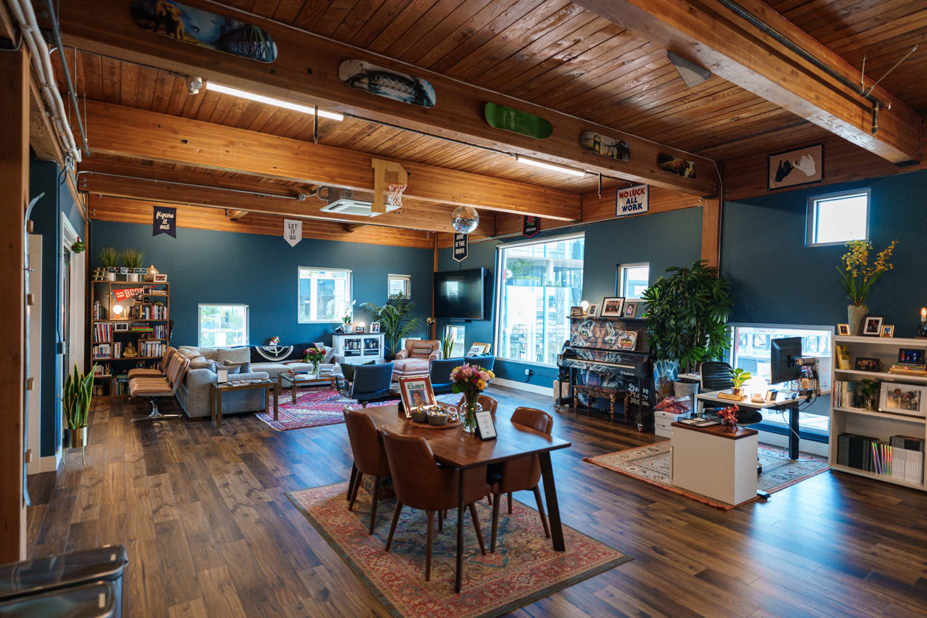 The image shows a spacious, modern office or communal area featuring wooden beams, a dining table, a lounge area with couches, and various decorative plants.