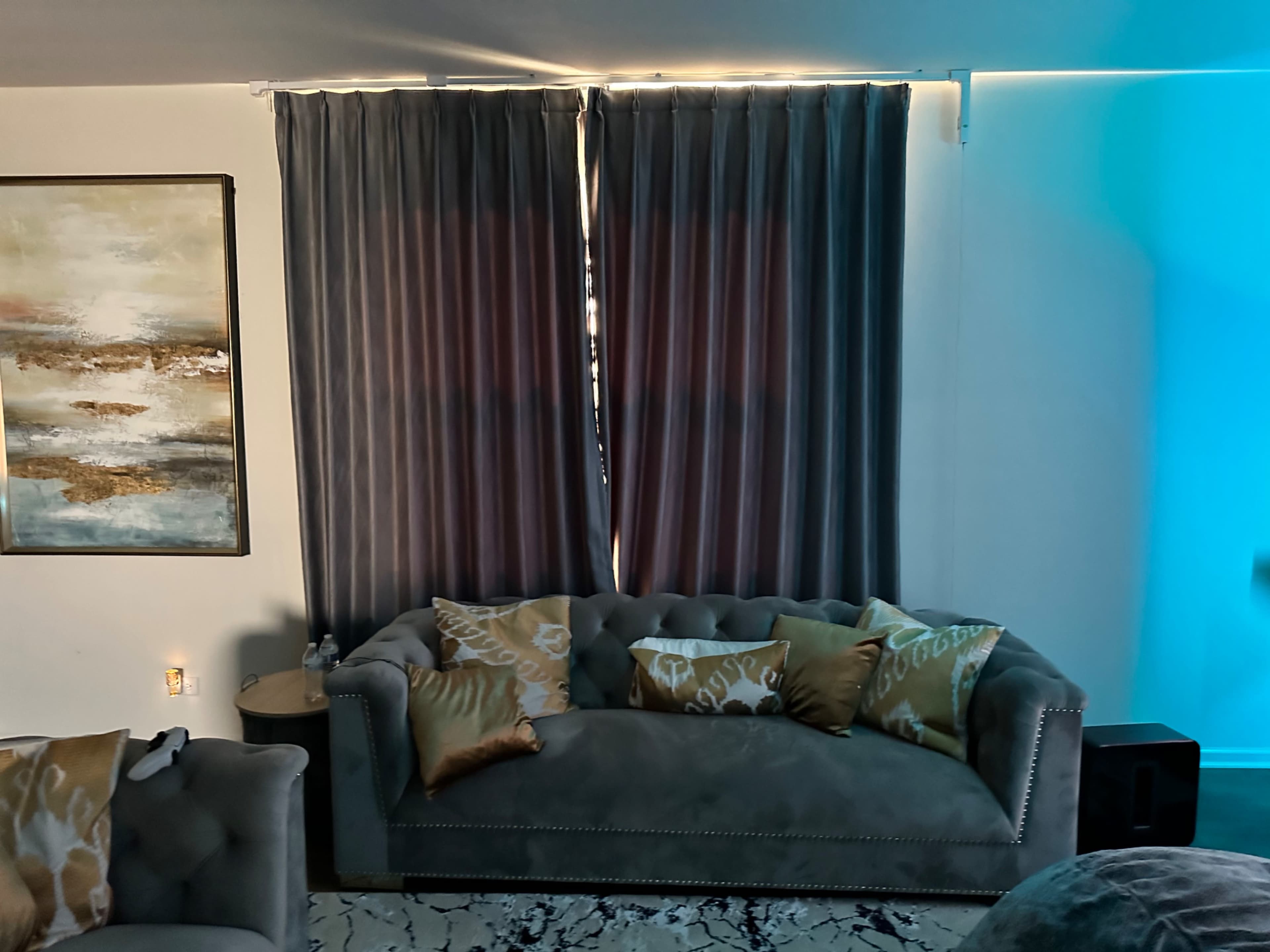 A living room corner featuring a dark gray sofa with patterned cushions, a large abstract painting on the wall, and curtains covering a window.