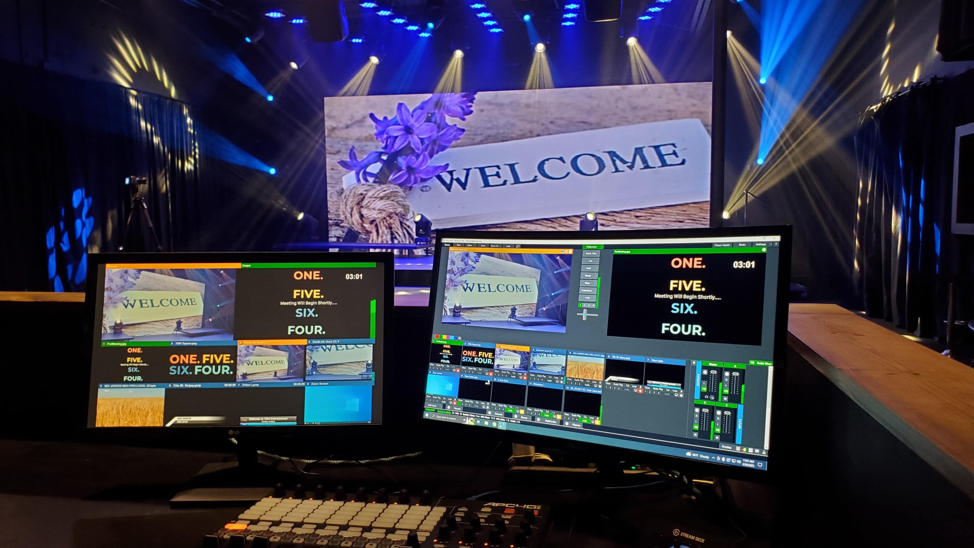 Two video monitors display on-screen graphics and text, while a large "WELCOME" sign is illuminated in the background, set on a stage with bright lighting.