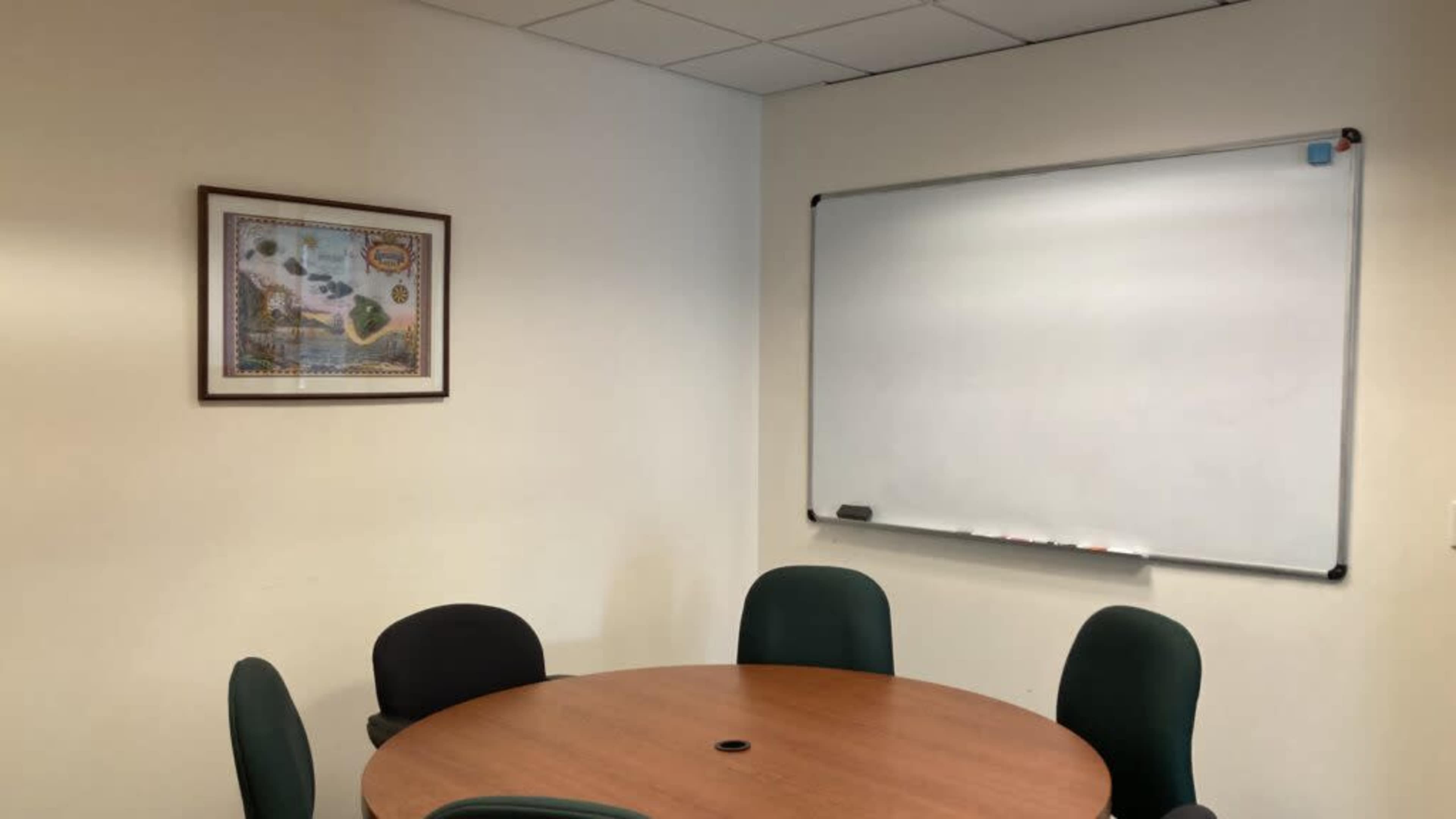 The image depicts a small conference room with a round wooden table surrounded by four green chairs, a whiteboard mounted on the wall, and a framed artwork on the adjacent wall.