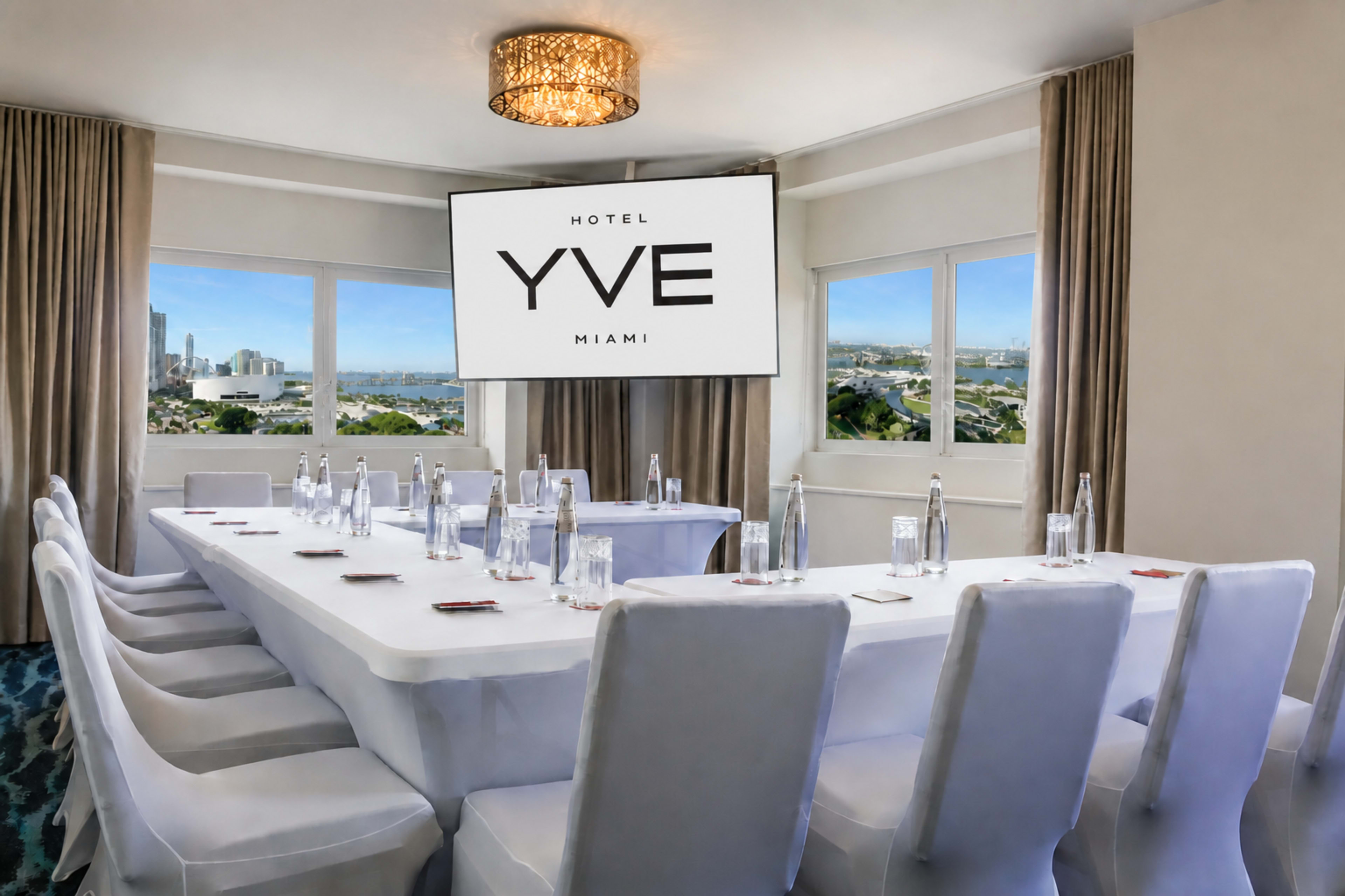 The image shows a conference room at Hotel YVE Miami, set up with a long table surrounded by chairs and a large screen displaying the hotel’s logo.
