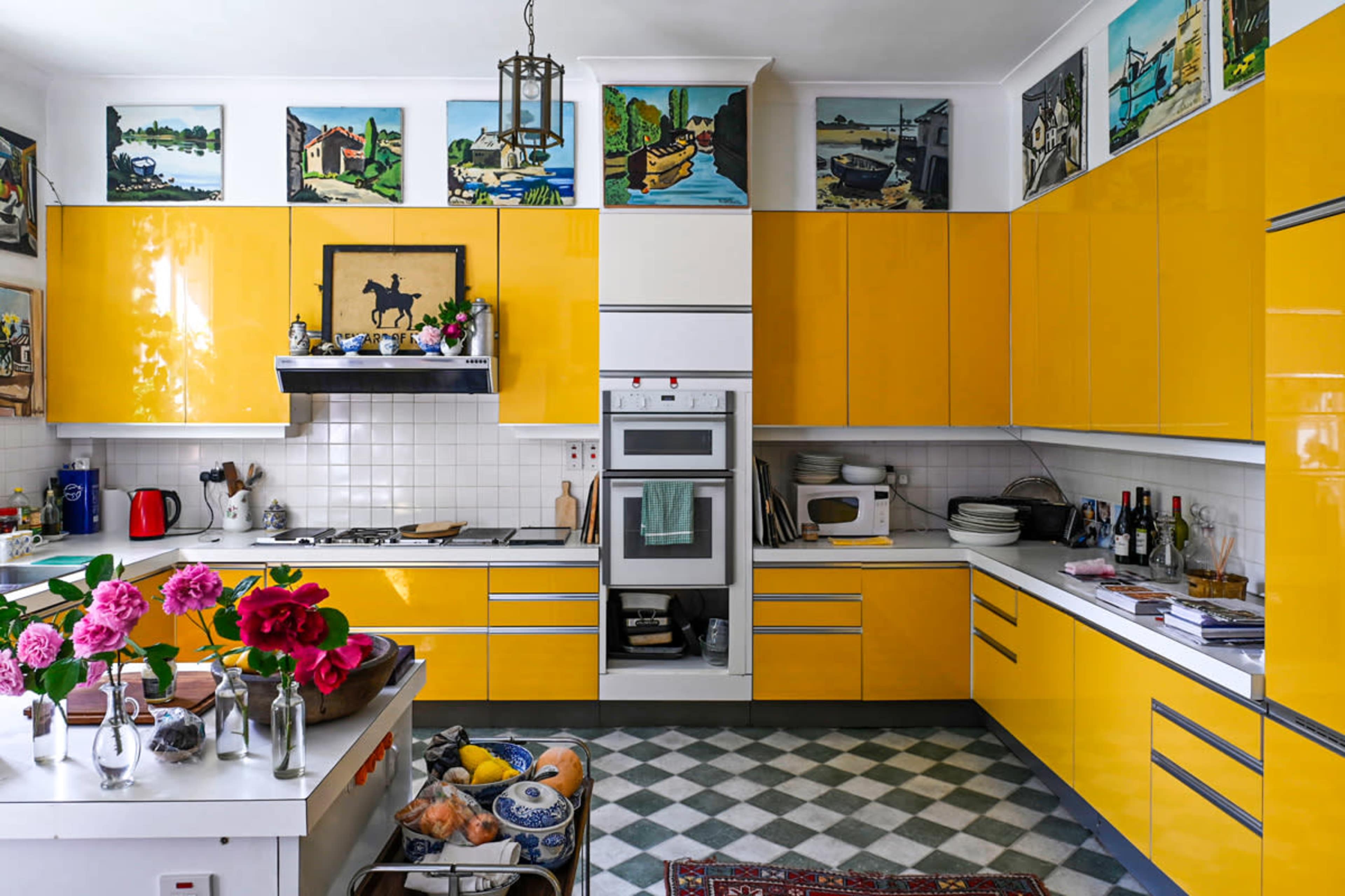 The kitchen features bright yellow cabinets, a checkerboard floor, and various paintings on the walls, with flowers and fruit displayed on the countertops.