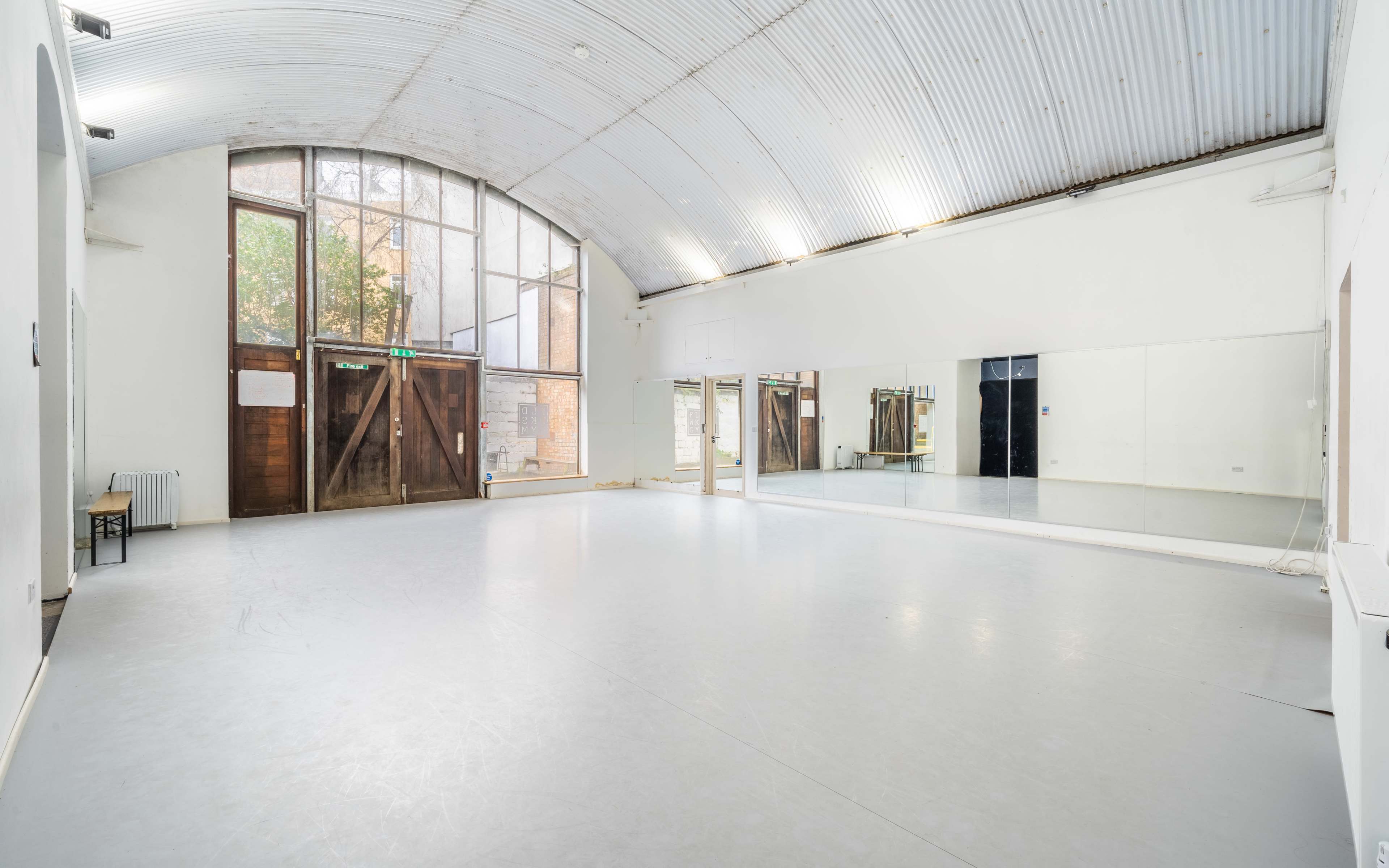 The image shows an empty dance studio with a high arched ceiling, large windows, and a mirrored wall.