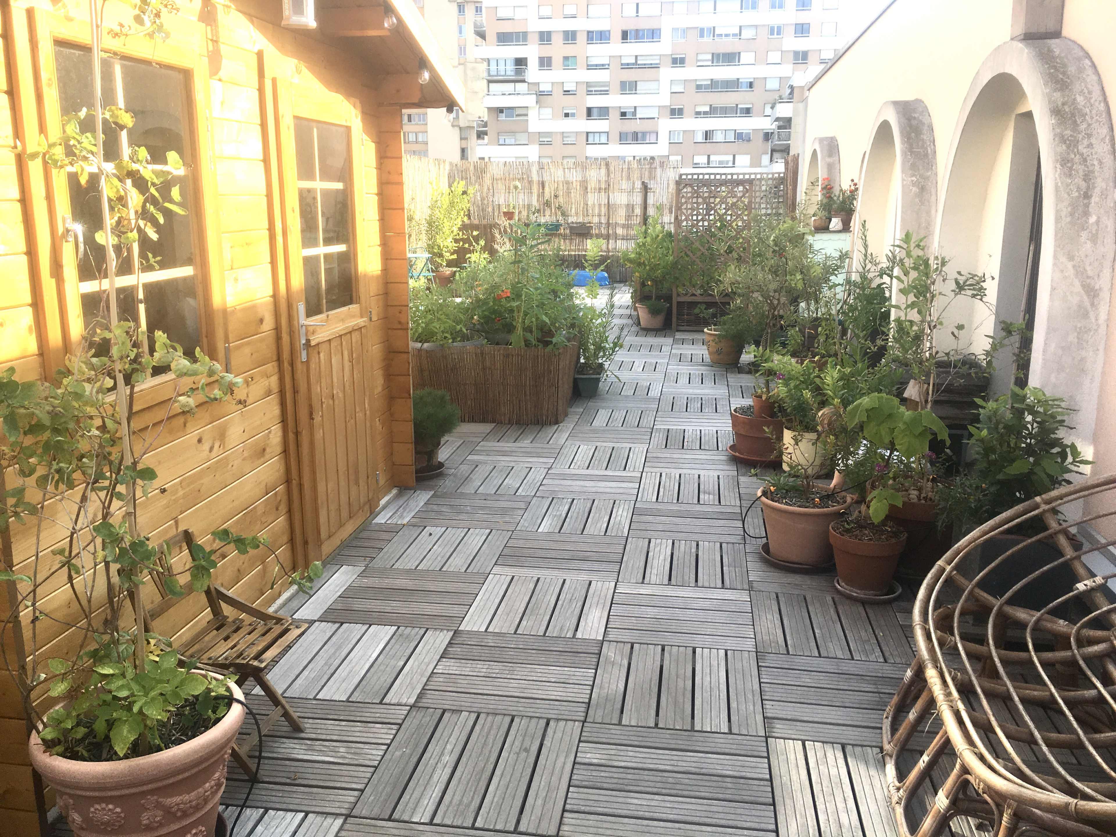 A wooden deck garden features potted plants and a small wooden shed, lined with tiles, in an urban setting.